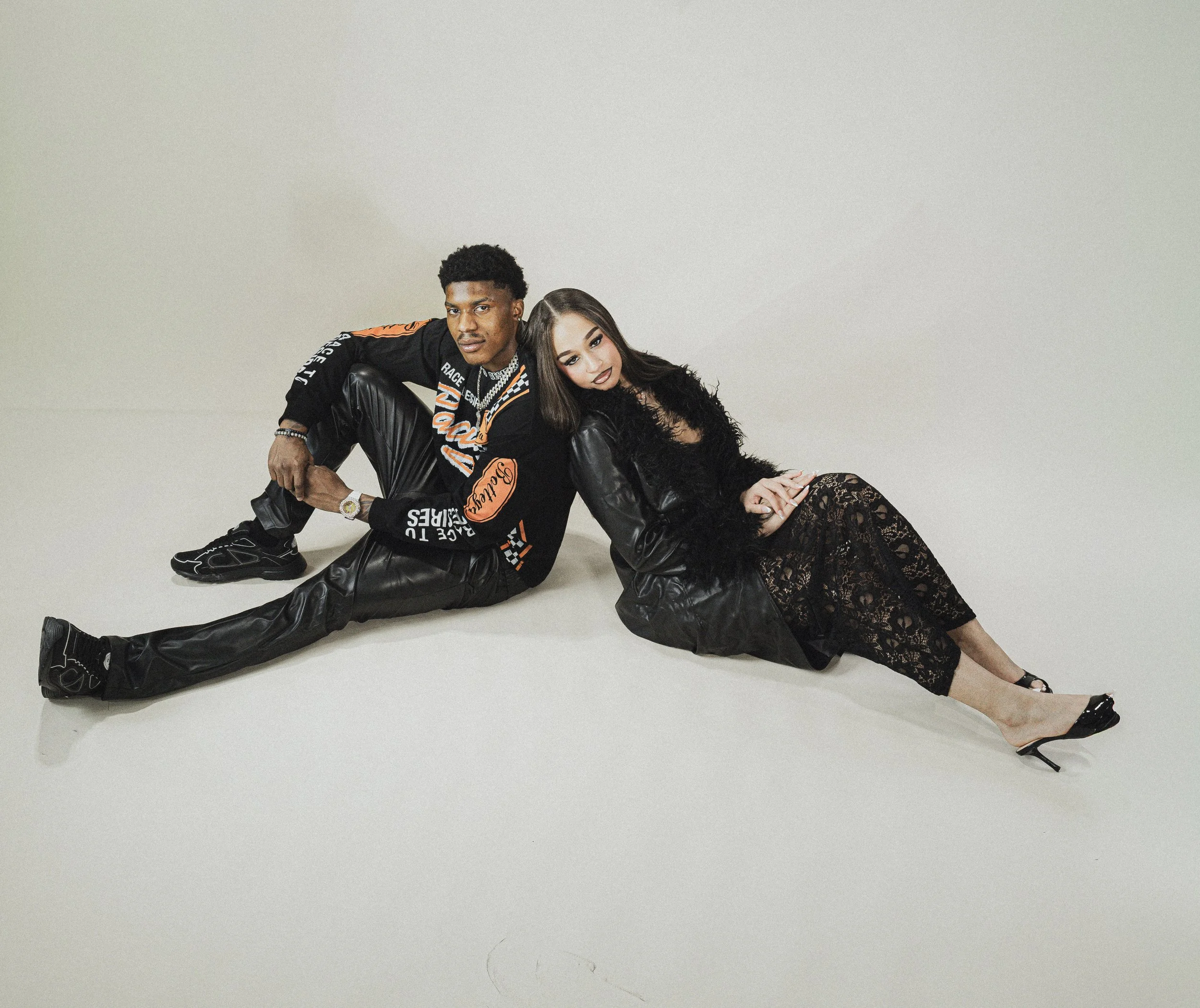 A young man and woman pose together on a plain white background. The man, with short curly hair, is dressed in a black and orange jacket, black leather pants, and sneakers. The woman, with long dark hair, wears lace black clothing and high heels. The