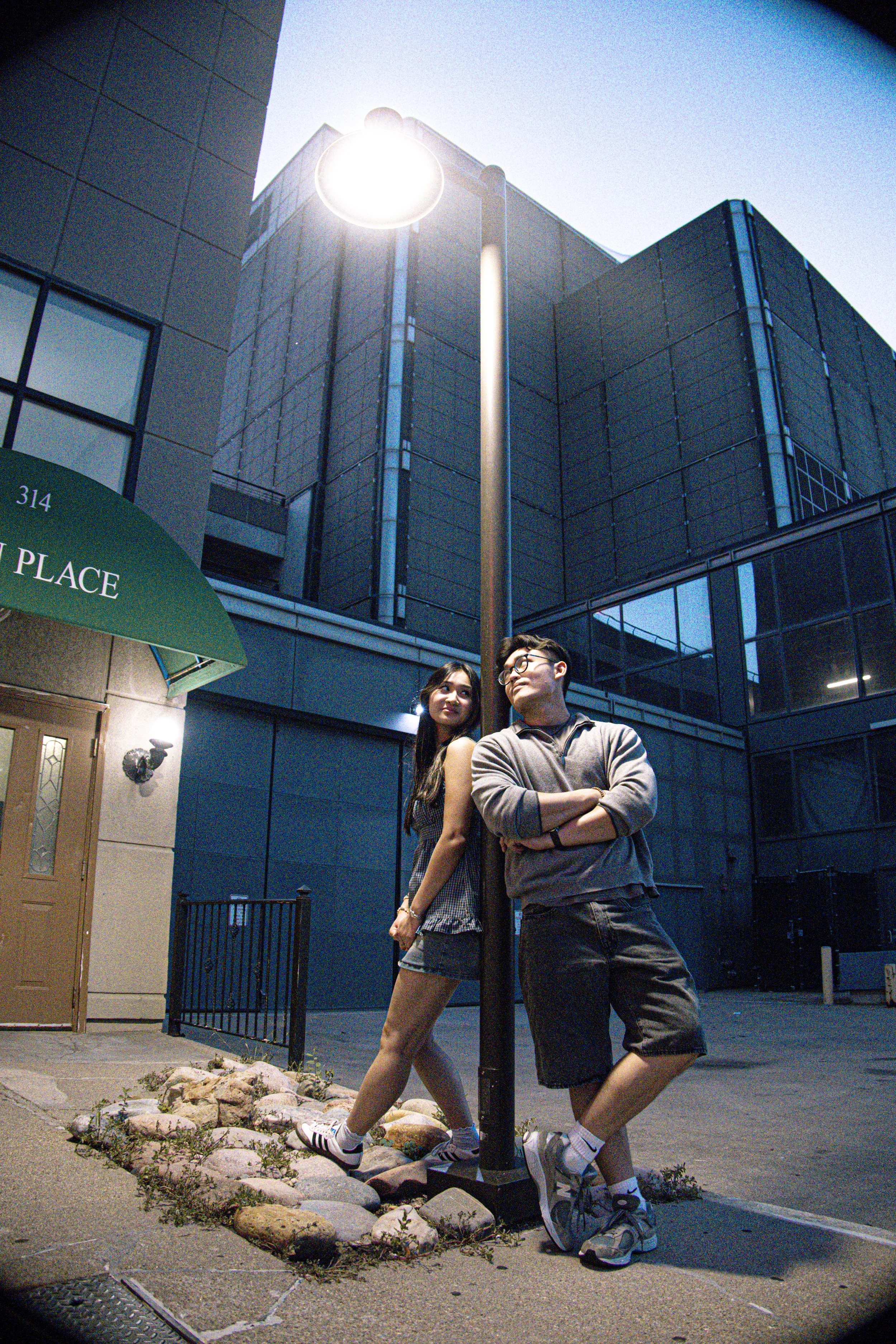 A young man and woman pose outdoors at night near a streetlamp, standing close with the woman leaning back against the pole and the man crossing his arms. They are in front of modern blue and gray buildings with a glass facade, and there are rocks an