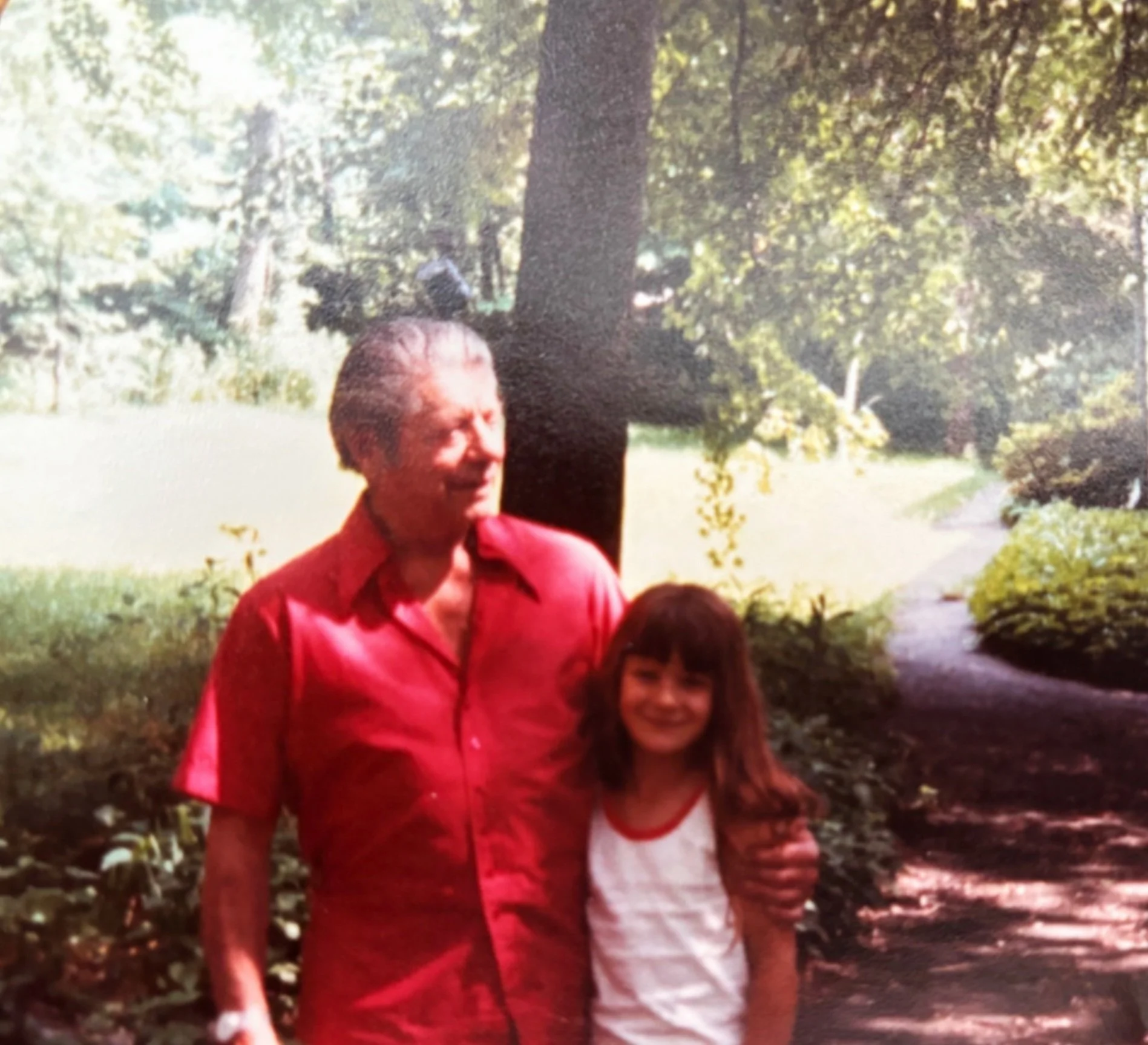 Seymour Fogel in a red jacket standing outdoors next to his granddaughter in a garden with leafy trees and green grass in the background.