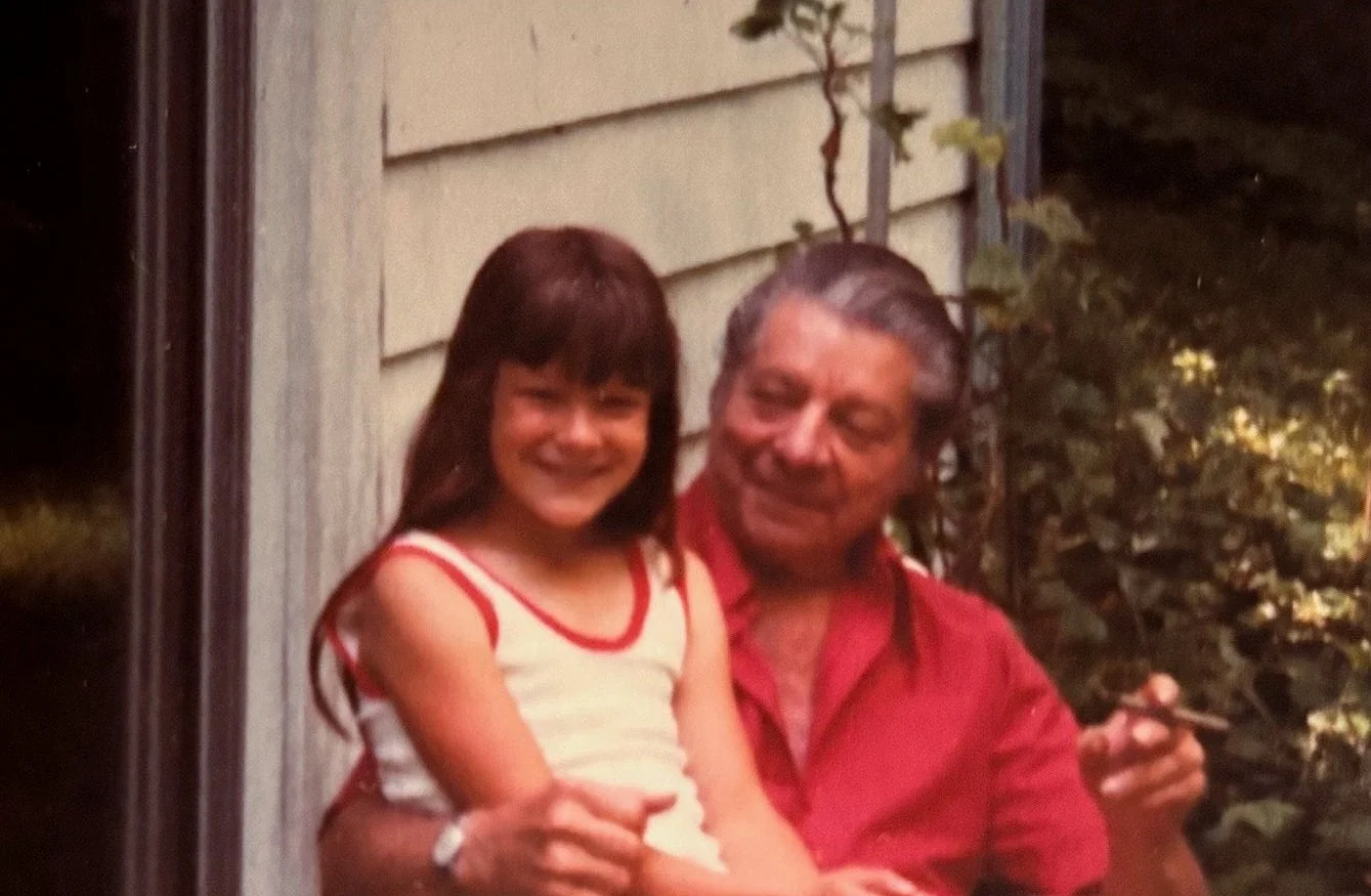 Seymour Fogel sitting with his granddaughter, smiling outdoors. She has brown hair and is wearing a white tank top with red trim. Fogel has gray hair and is wearing a red shirt, holding a cigarette.