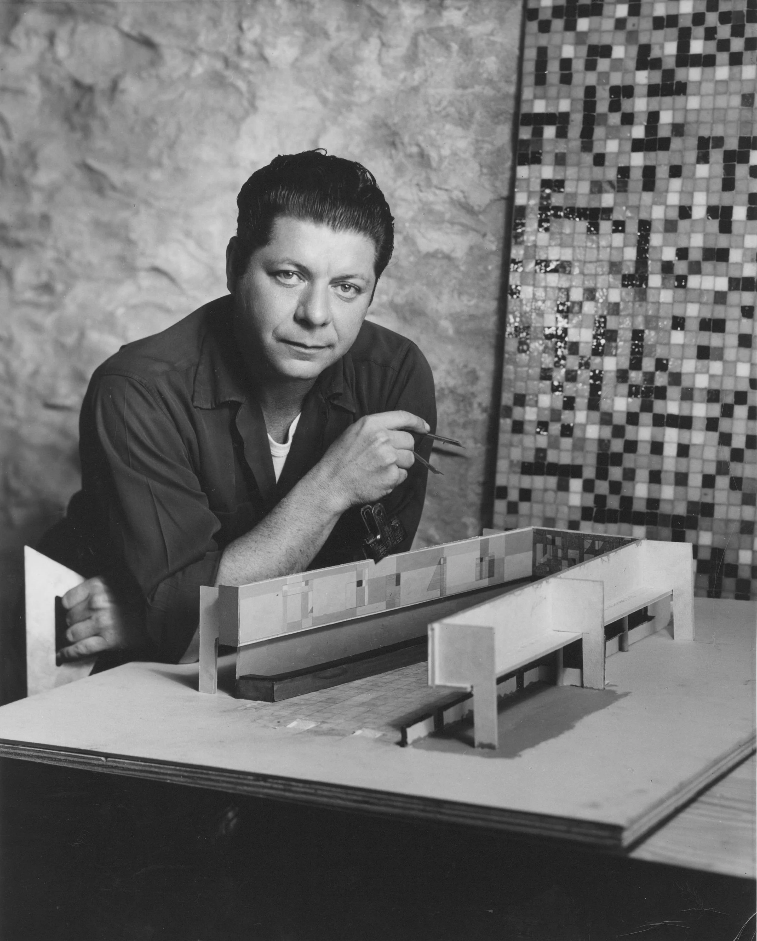 Black and white photo of Seymour Fogel sitting at a table with a miniature architectural model, against a stone wall and a tiled mosaic background.