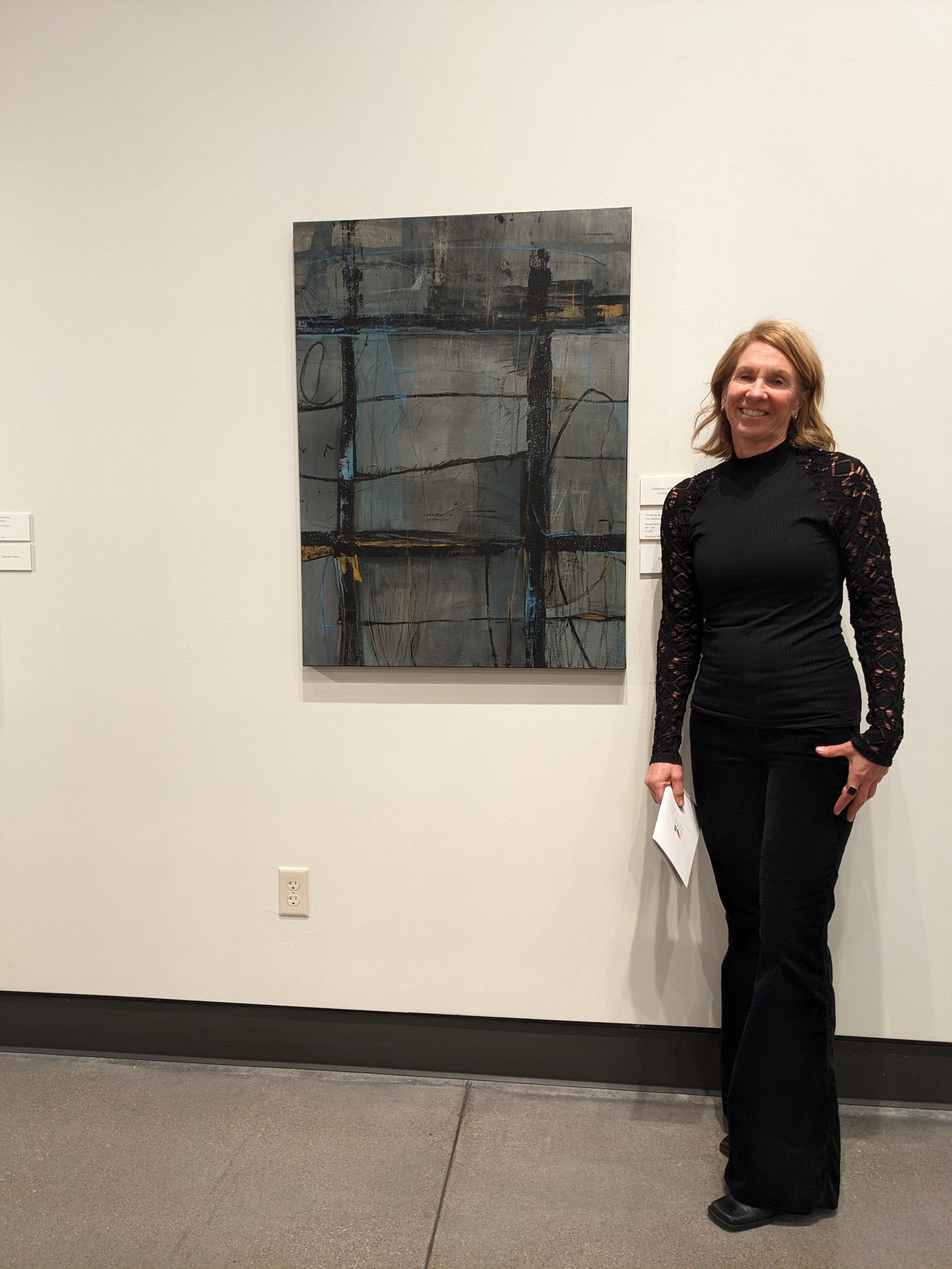 Christine wearing a black lace long-sleeve top and black pants standing beside an abstract painting at an art gallery.