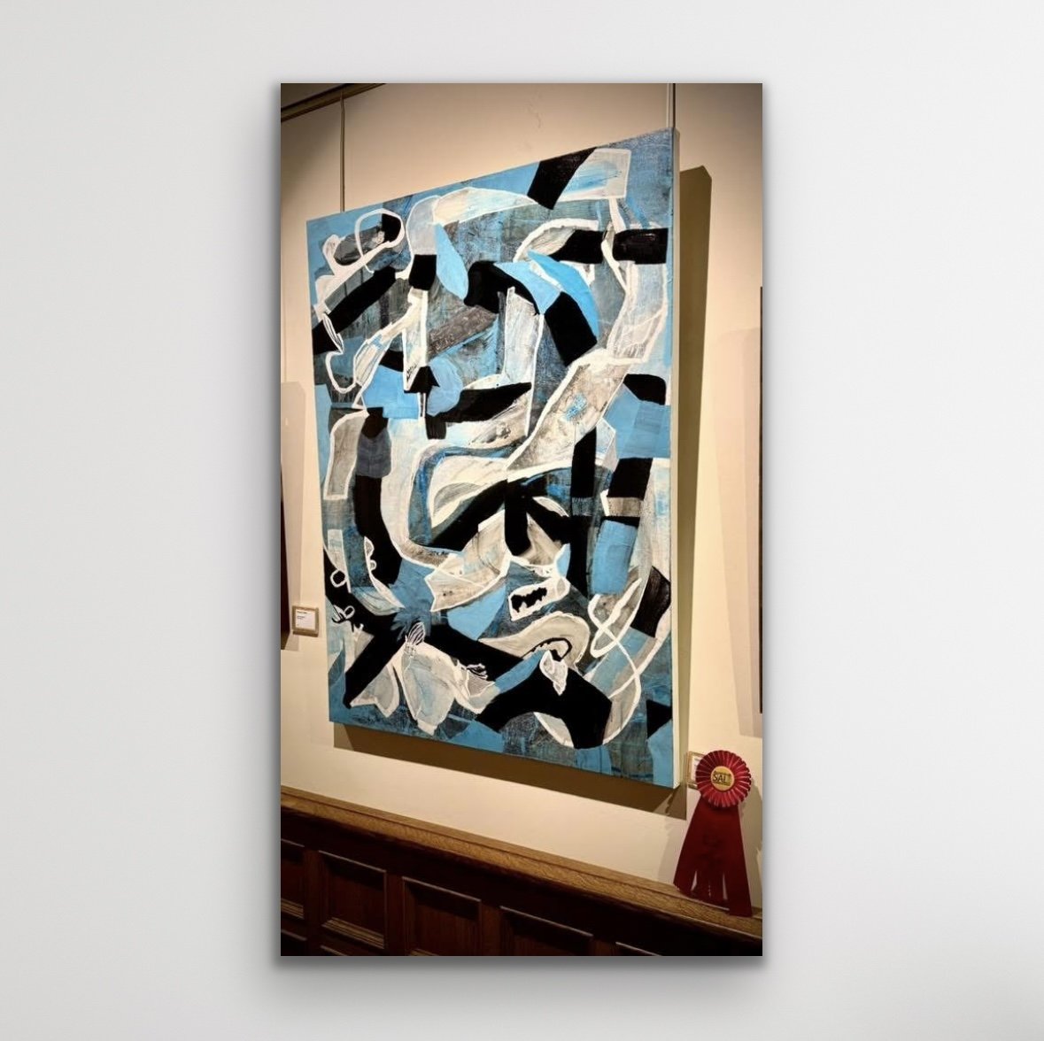 Abstract blue, black, white, and gray painting displayed on a wall, with a red ribbon award placed below it.