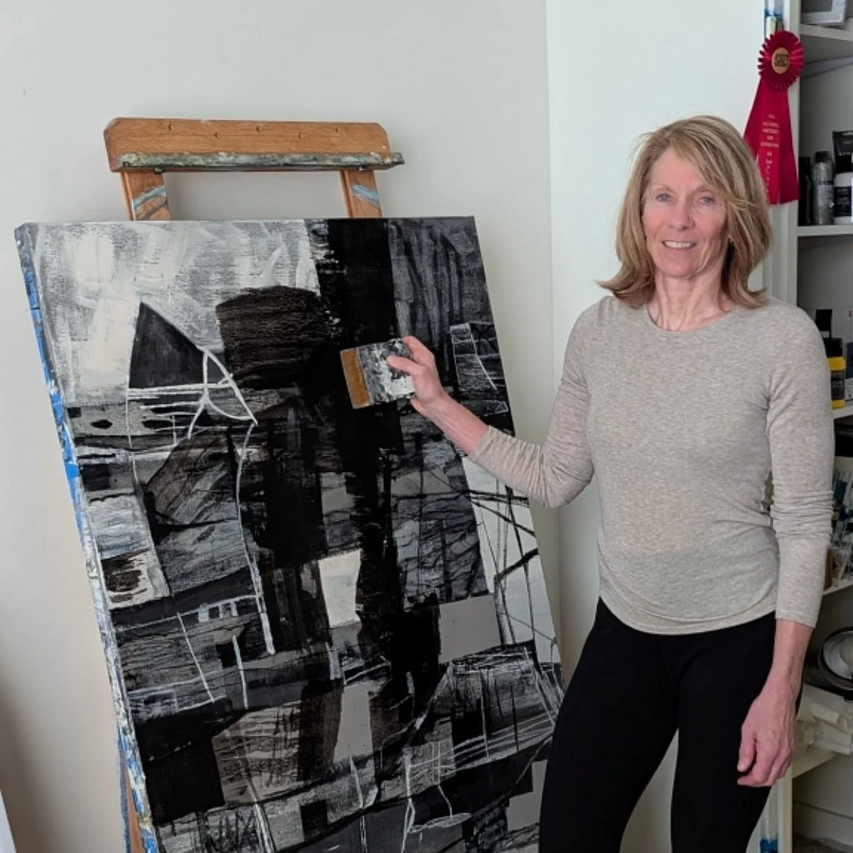Christine in a gray long sleeve shirt and black pants painting an abstract black and white artwork on a large canvas in a room with a bookshelf and a red ribbon award hanging on the wall.