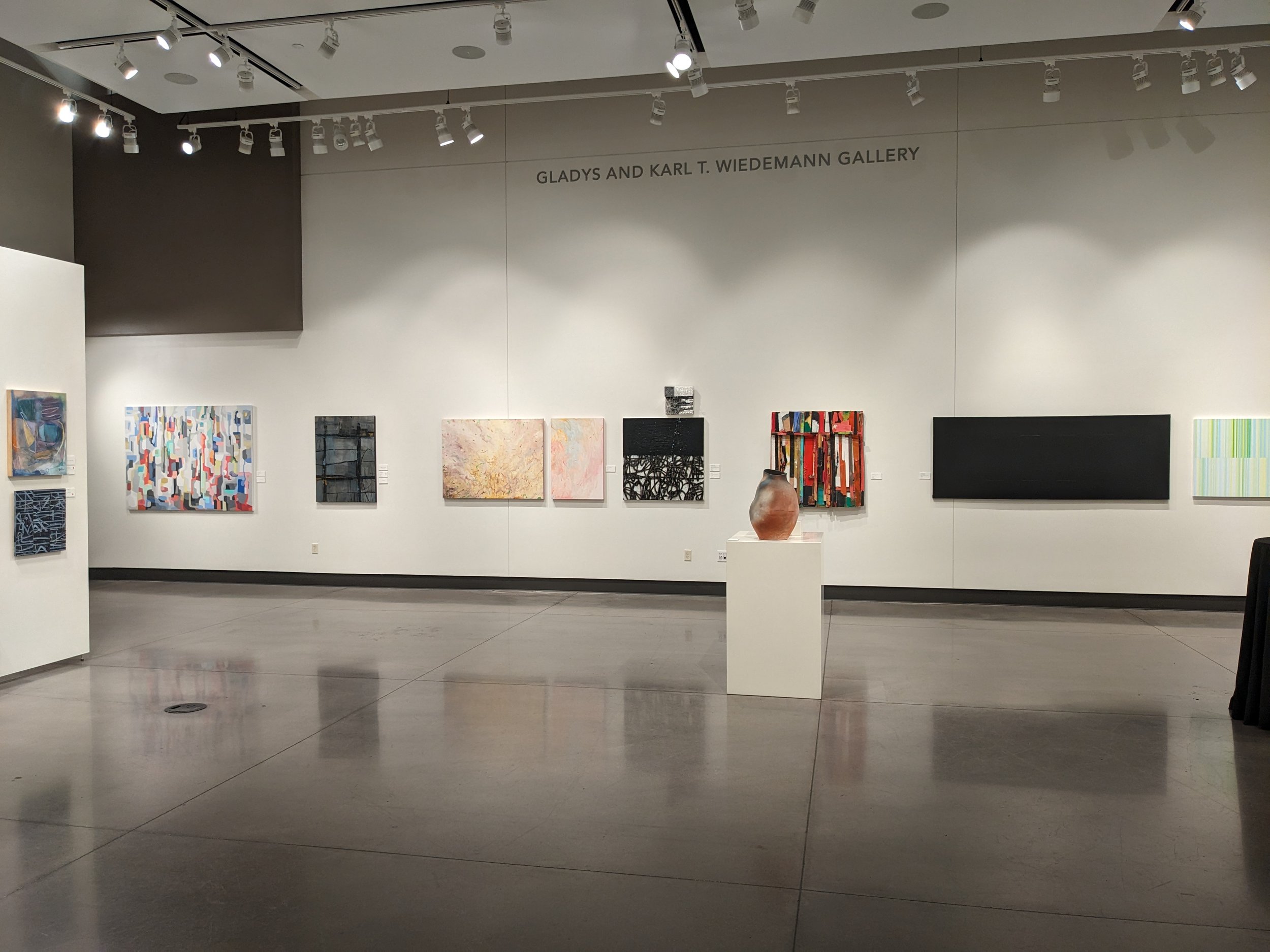 Interior of an Mark Arts gallery with various abstract paintings on the walls and a ceramic vase on a pedestal.
