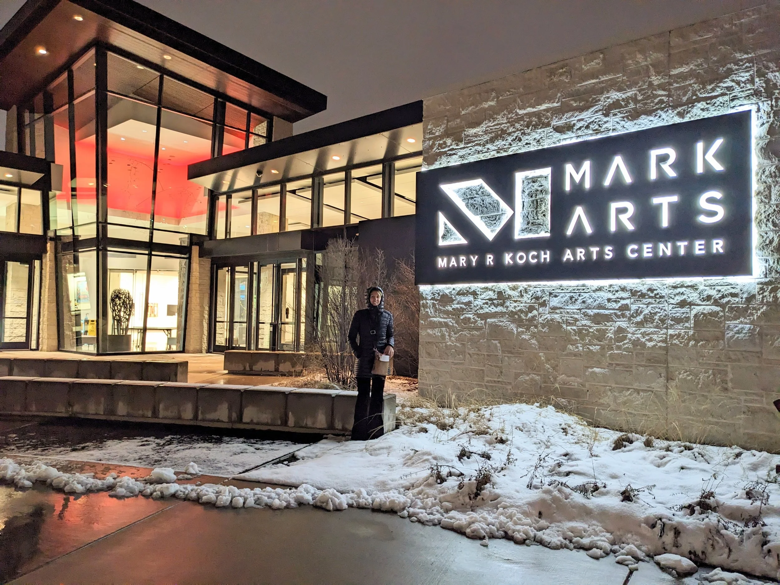 Exterior night view of the Mary R. Koch Arts Center with a lit sign that reads 'Mark Arts Mary R. Koch Arts Center', snow on the ground, a person standing outside, and the building's modern glass and brick architecture.
