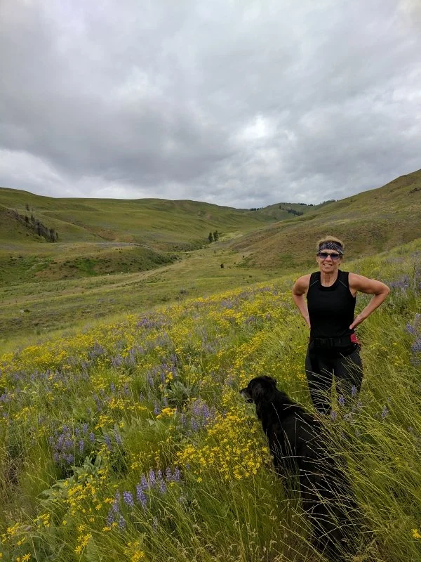 A woman with short hair, wearing sunglasses and a black athletic outfit, standing in a meadow with wildflowers, smiling with her hands on her hips, accompanied by a black dog, in a hilly green landscape under an overcast sky.