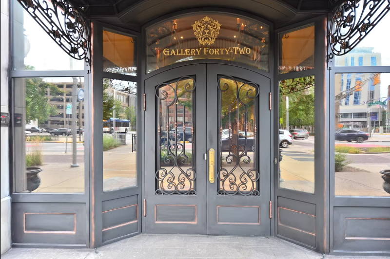 The entrance of Gallery Forty-Two, featuring double doors with ornate wrought iron designs, glass panels, and a gold-colored handle. Above the doors, a sign reads 'Gallery Forty-Two' in gold lettering, framed by decorative metalwork.