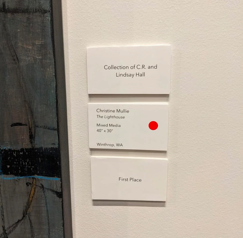 A sign on a white wall with information about an art piece titled 'The Lighthouse' by Christine Mullie, measuring 40 by 30 inches, from Winthrop, Washington, and awarded first place. The sign has a red dot indicating it is sold