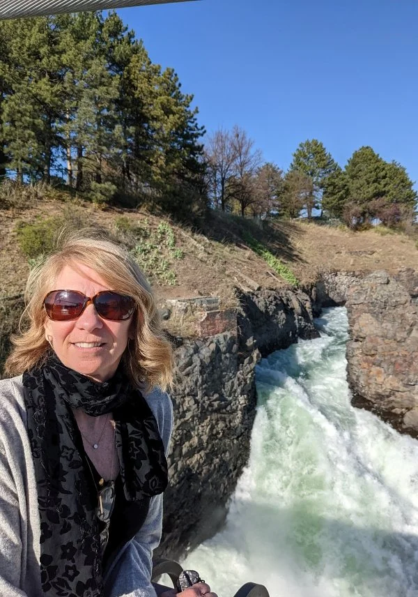 A woman with blonde hair wearing sunglasses, a black scarf, and a gray jacket smiling outdoors near a flowing river with rocky cliffs and green trees under a clear blue sky.