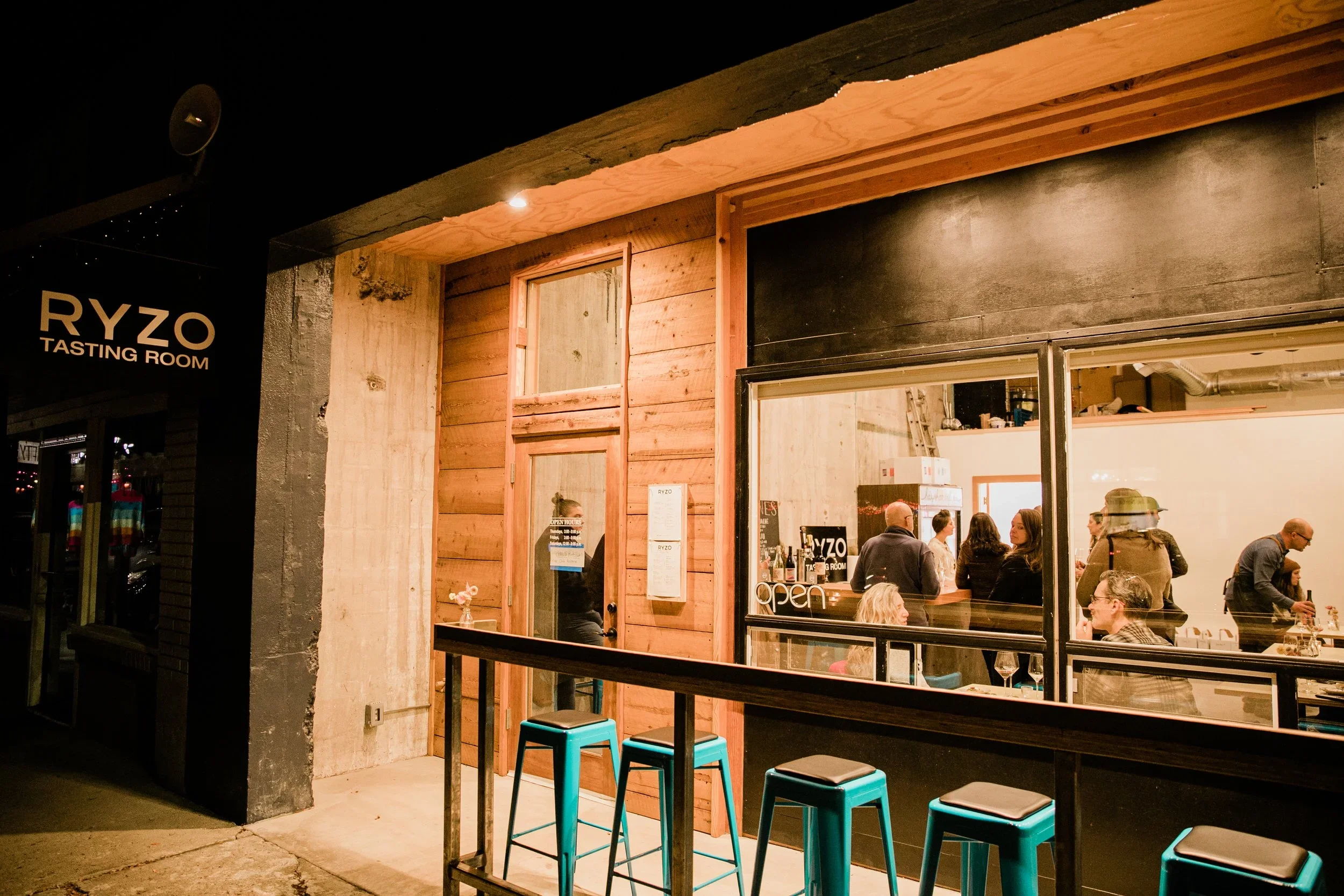 Night view of a restaurant named Ryzo Tasting Room with a large window showing people inside; outdoor seating with teal stools; wooden and concrete facade