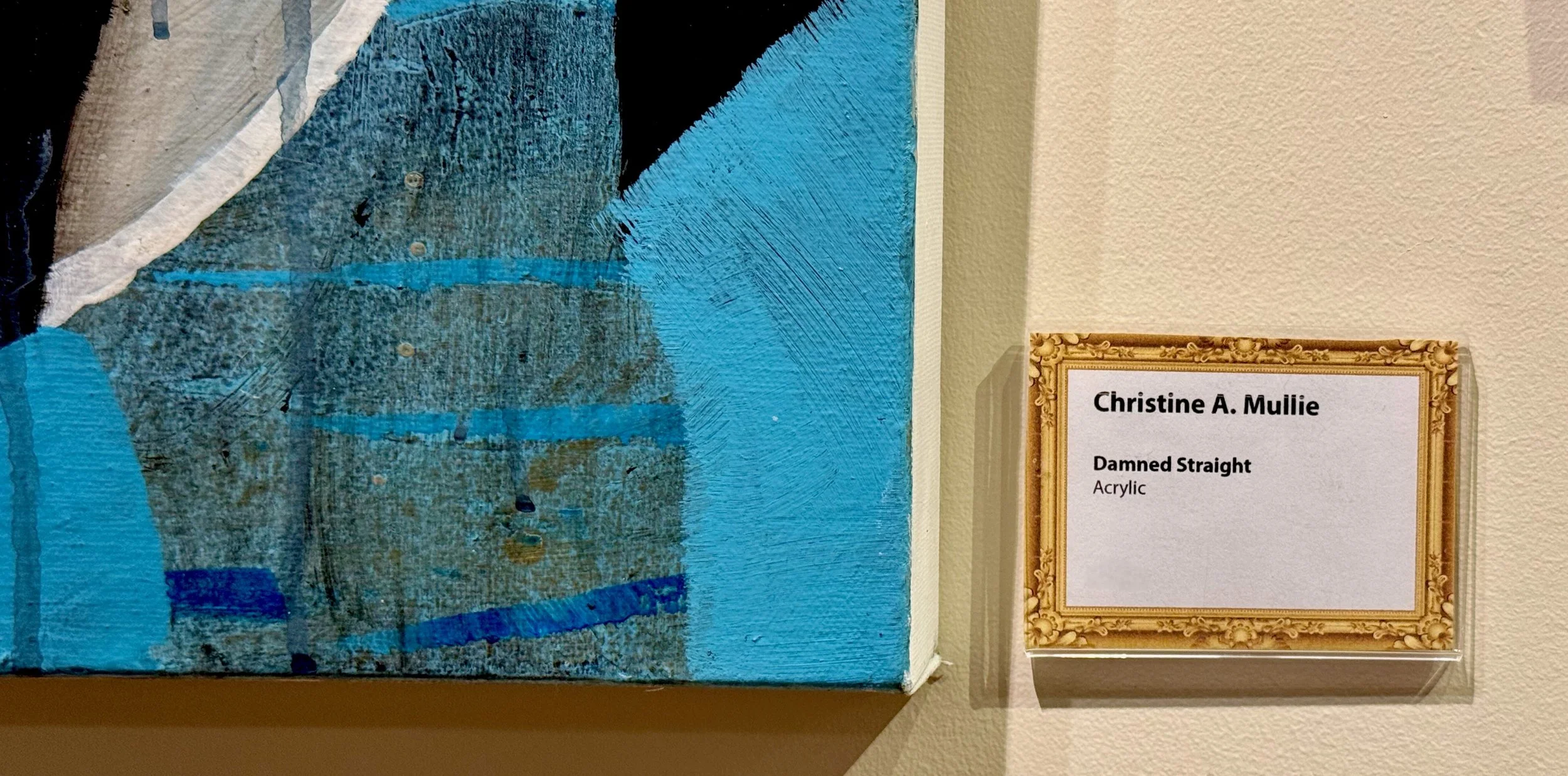 Close-up of an abstract acrylic painting with blue, gray, black, and white brushstrokes, alongside a label that reads 'Christine A. Mullie, Damned Straight, Acrylic.'