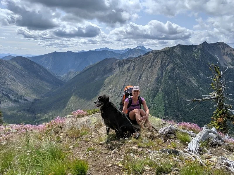 A woman with a backpack sitting on a mountain trail beside a black dog, overlooking a valley and mountain range with cloudy sky.