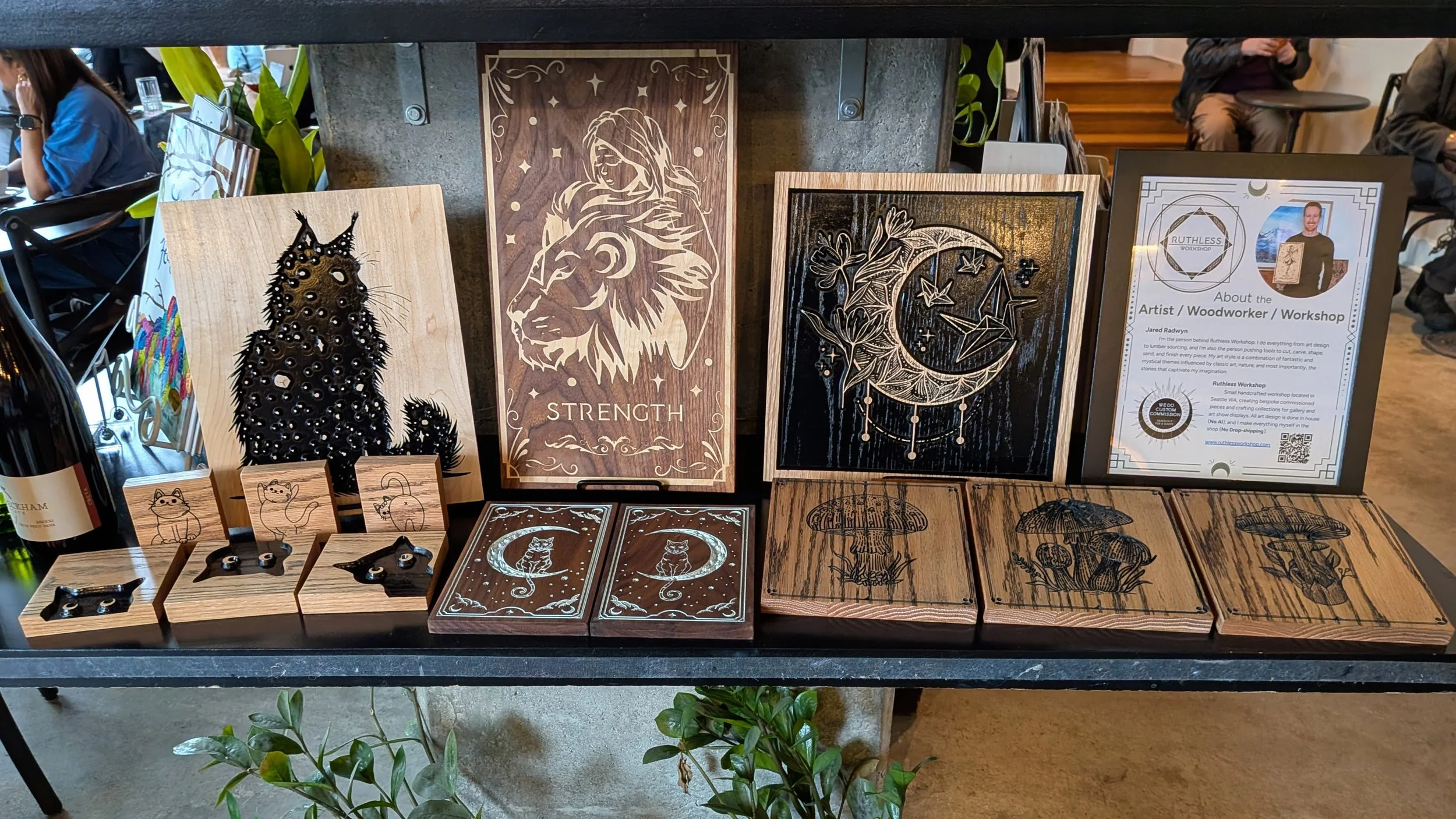 Display of handcrafted wooden artwork featuring mystical and animal designs, including a cat, lion, and moon with stars, along with informational sign about the artist and workshop.