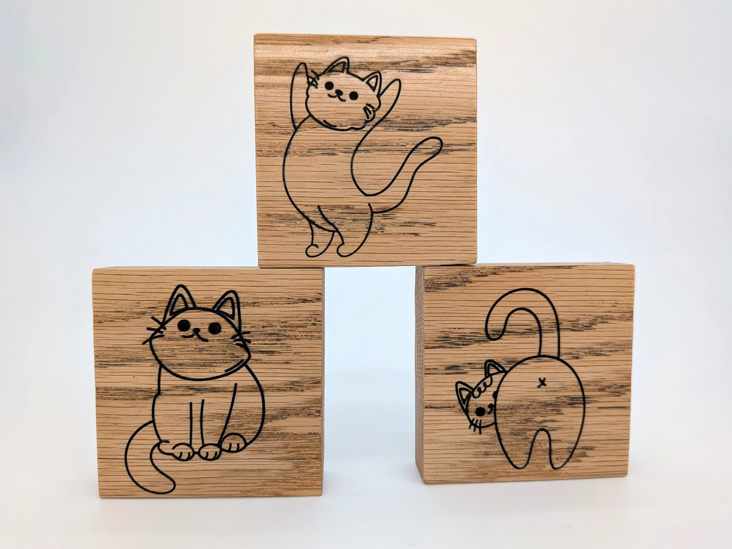 Three handcrafted wooden blocks with carved black lines of cats. The top block shows a cat stretching with paws up; the bottom left block shows a sitting cat with a curled tail; the bottom right block shows a back view of a cat with a raised tail.