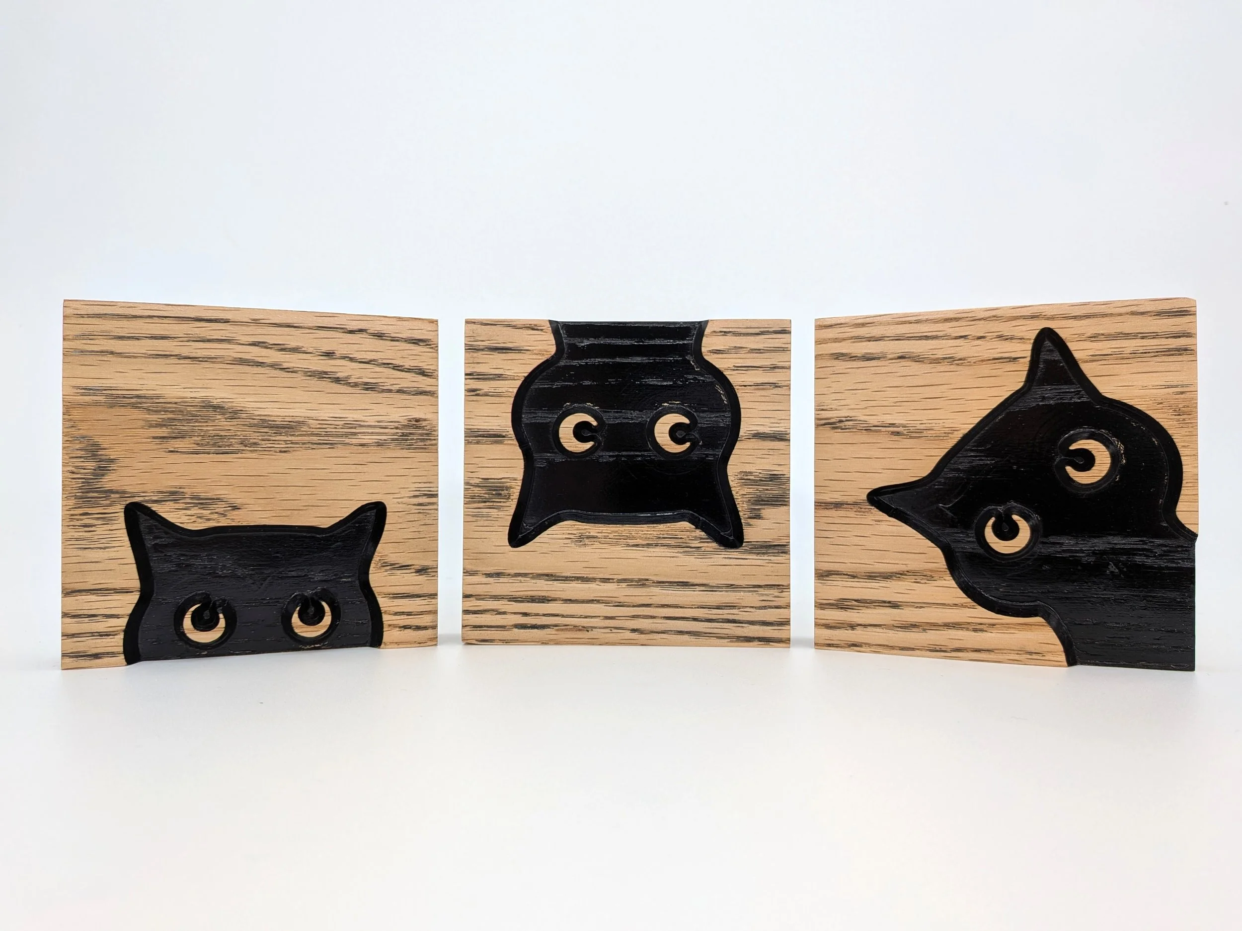Three handcrafted wooden block carvings of black cat-shapes peeking out, arranged in a row on a white surface.