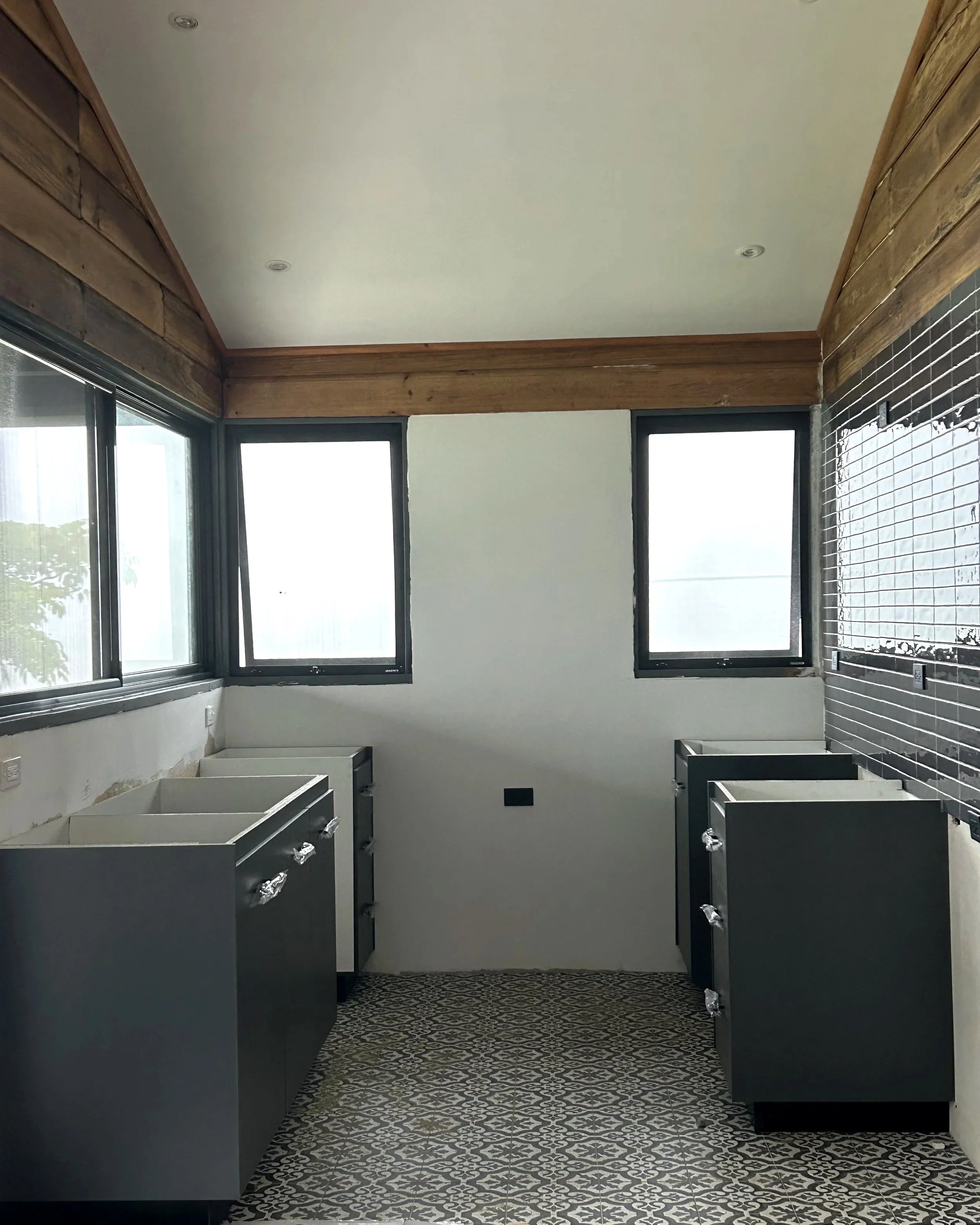 Interior de una habitación en construcción con ventanas grandes, suelos de azulejo en patrón geométrico, paredes blancas y acabados en madera.