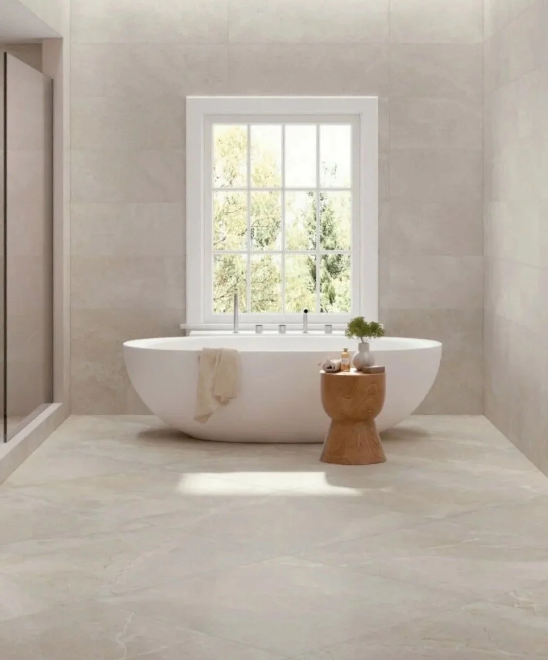 Un baño con una bañera ovalada blanca junto a una ventana grande, con vistas a árboles. En una esquina hay una mesa pequeña de madera con una planta y algunos objetos decorativos. La decoración es minimalista y luminosa.