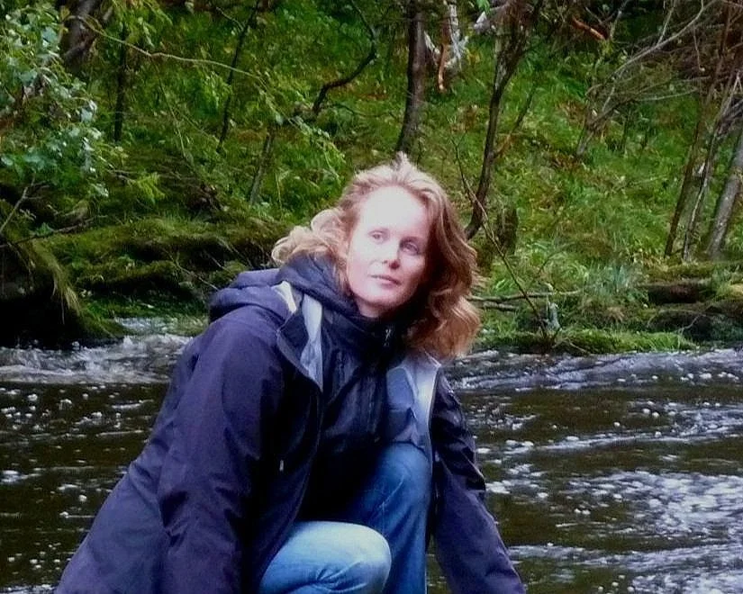 Junge Frau in dunkler Jacke und blauer Jeans sitzt bei flachem Fluss im Grünen.