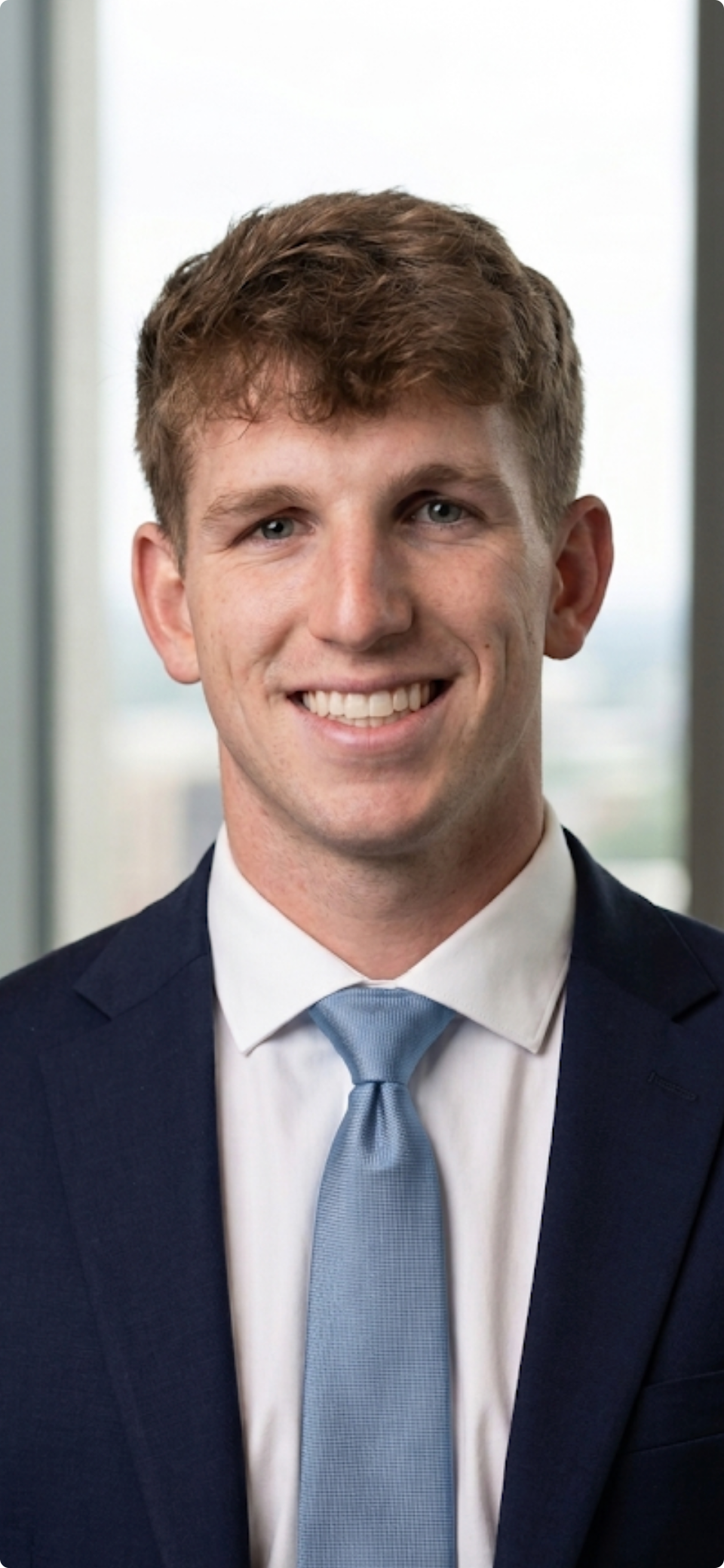 Portrait of a young man with brown hair, blue eyes, wearing a dark suit, white shirt, and light blue tie, smiling in front of a window.