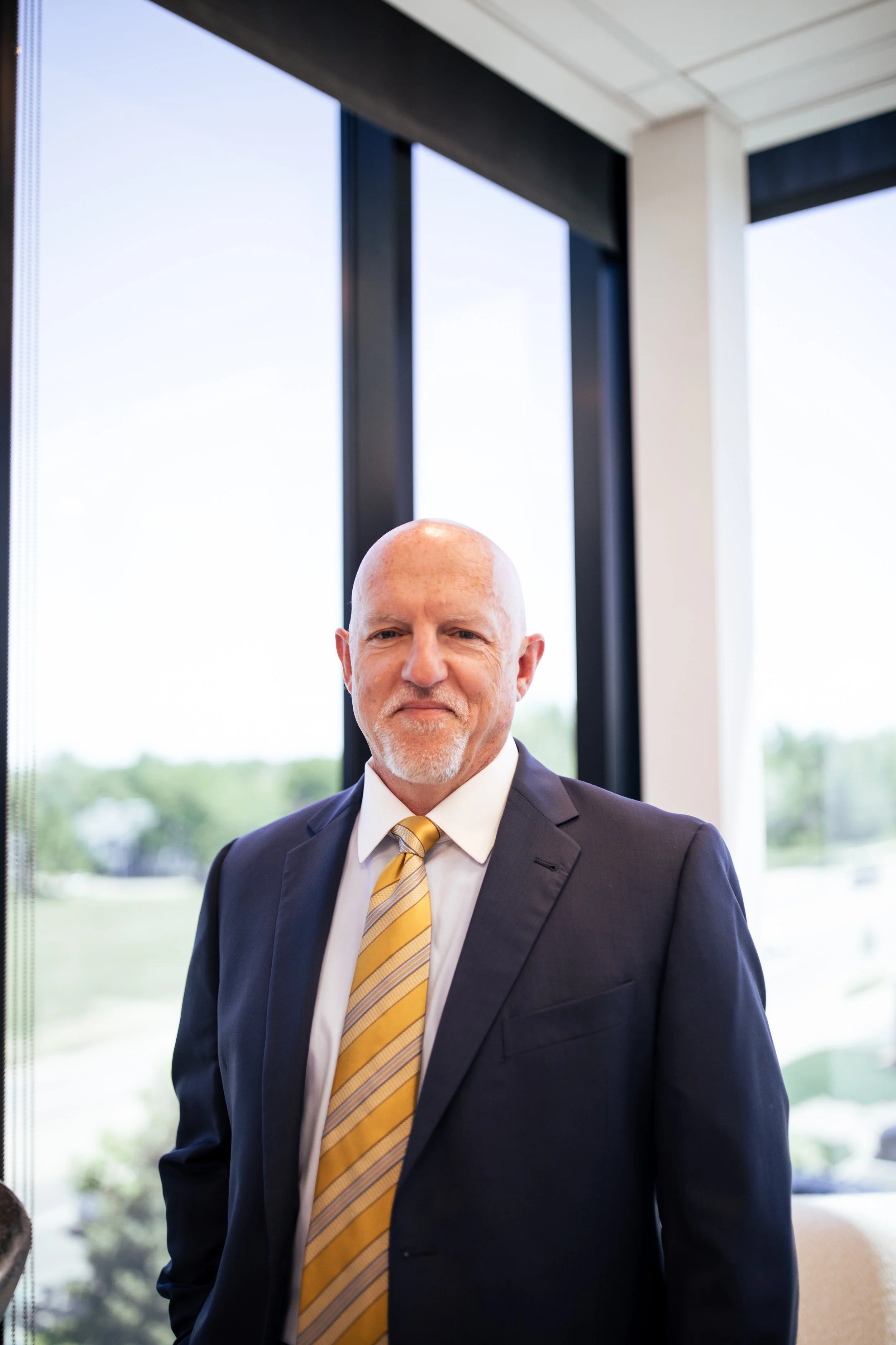 A mature man with a bald head and a white beard, wearing a dark suit, white shirt, and a yellow striped tie, standing indoors near large windows with a view of greenery outside.