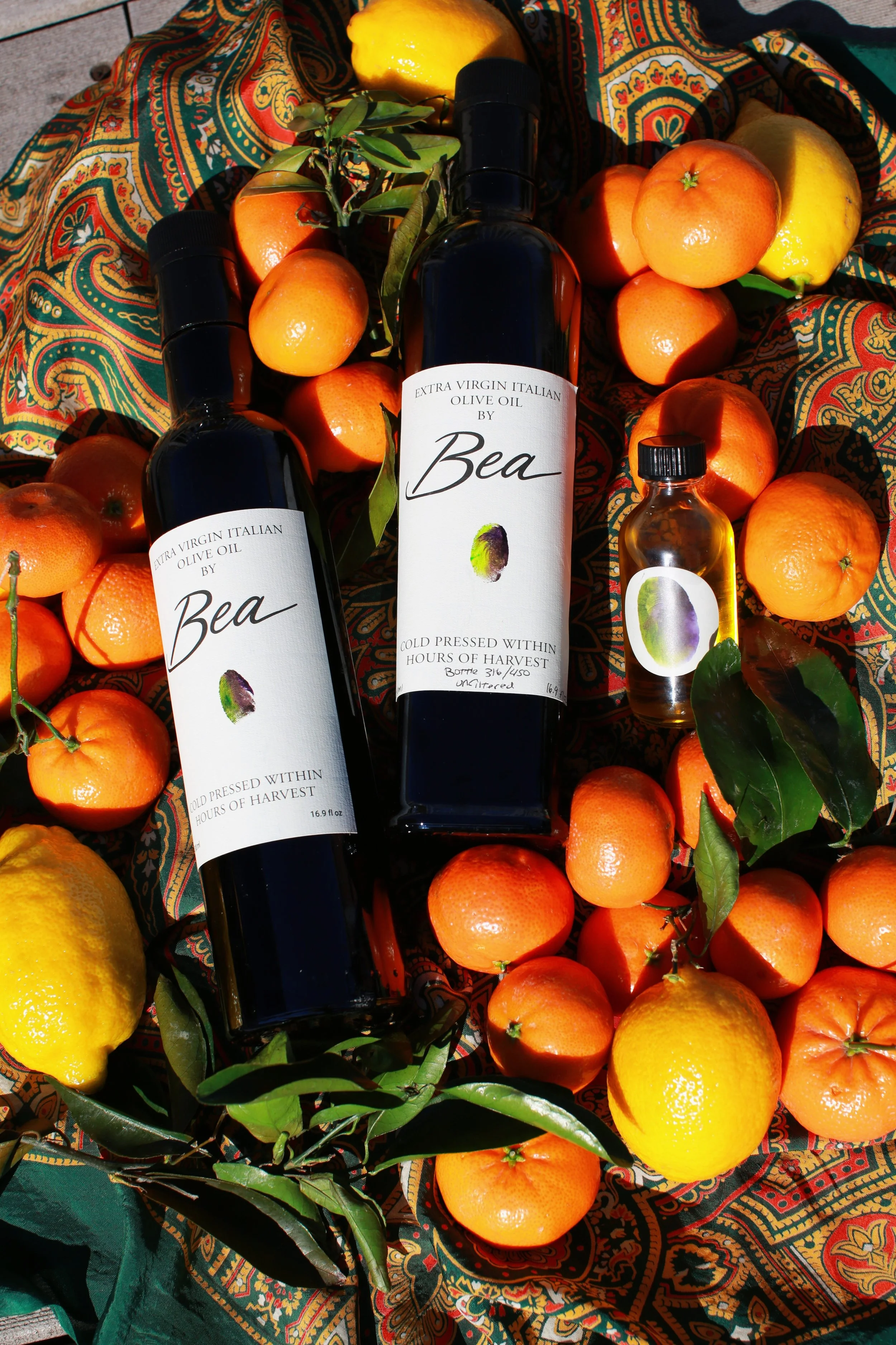 Two bottles of Bea extra virgin Italian olive oil surrounded by oranges, lemons, and green leaves on a patterned cloth.