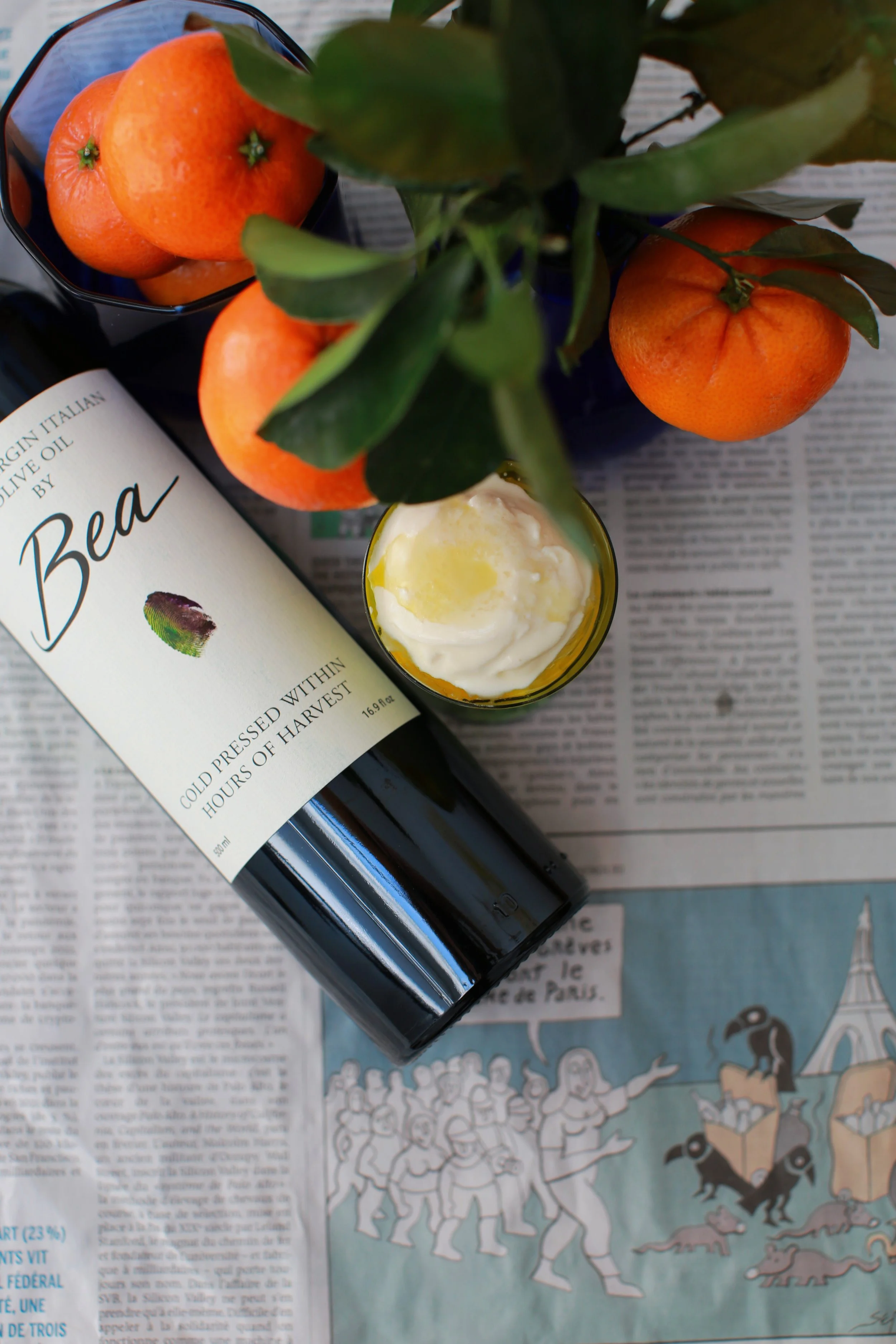 A bottle of olive oil labeled 'Bean' lying on a newspaper, with a glass of yogurt, a bowl of oranges, and a leafy plant nearby.