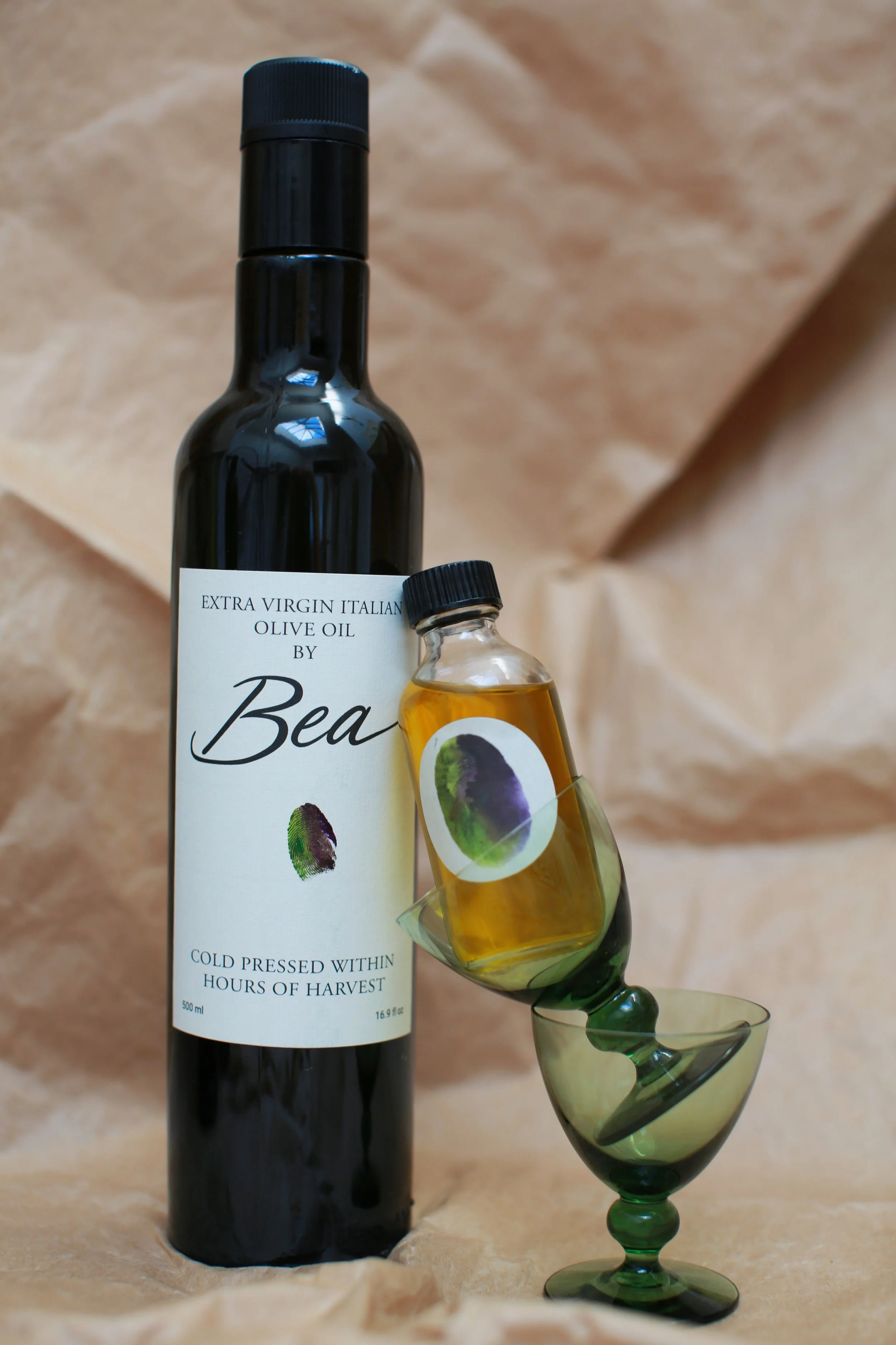 A black bottle of extra virgin Italian olive oil labeled 'Bea' with a small glass bottle of olive oil stacked on a green glass goblet, all set against a beige textured background.