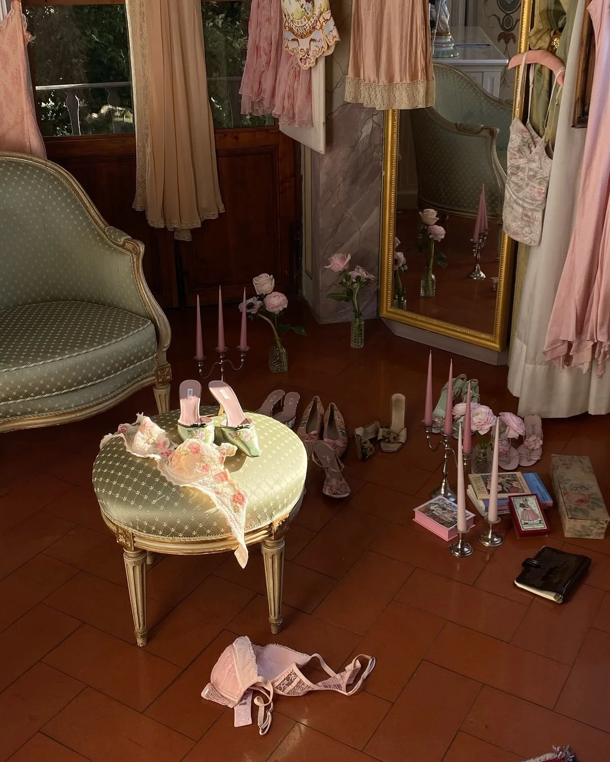 Una habitación decorada en estilo vintage con un sillón verde claro, un puff a juego, varios pares de zapatos, una balda con libros, candelabros con velas rosas, flores en jarrones, ropa y accesorios de moda, y un espejo de piso.