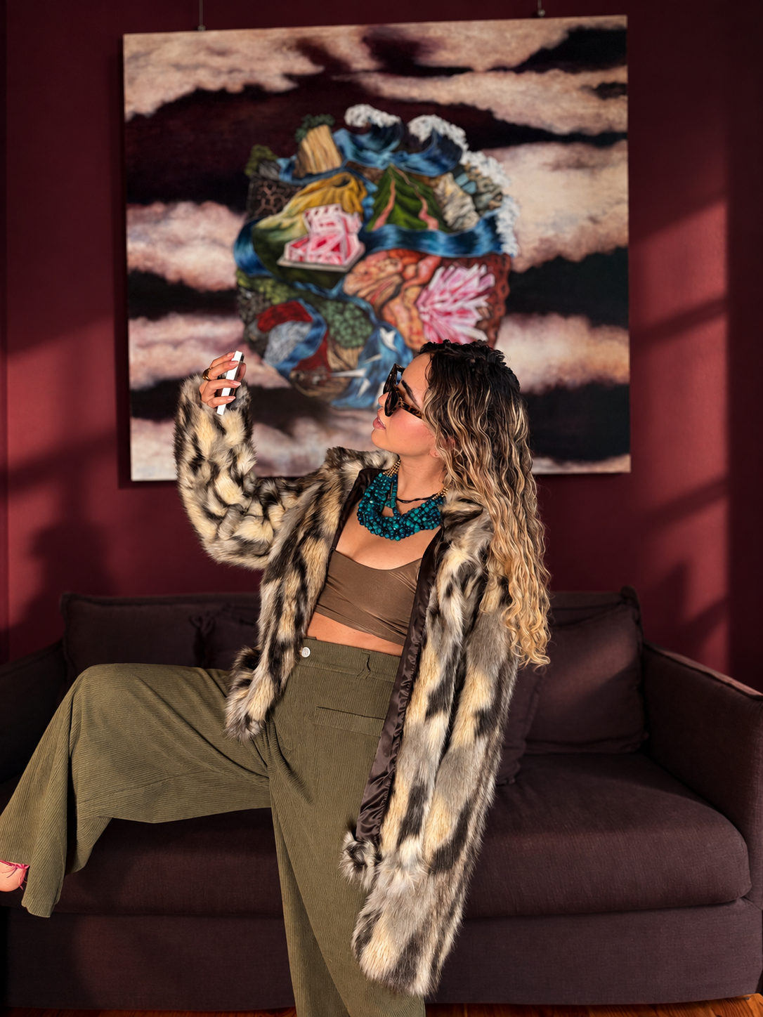 Mujer con cabello rizado y gafas de sol tomando una selfie, vestida con abrigo de piel y pantalones anchos en un sillón, con un cuadro abstracto en la pared de fondo.