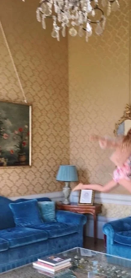 Una niña saltando en una sala elegantemente decorada con paredes doradas, decoraciones doradas, un candelabro de cristal, un sofá azul, una lámpara con pantalla azul y un cuadro en la pared.