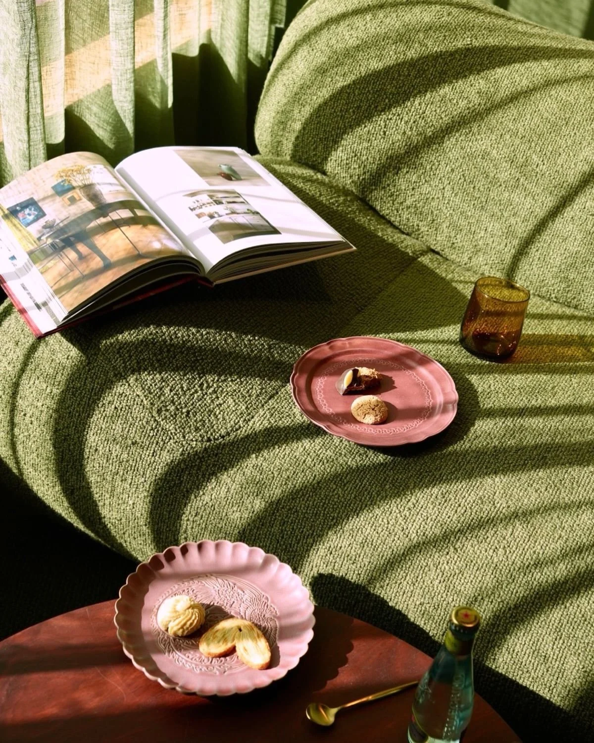 Sofá de color verde con una revista abierta y una taza de vidrio en una mesa, también hay un plato rosa con galletas y chocolates. Luz solar. Cortinas verdes al fondo.