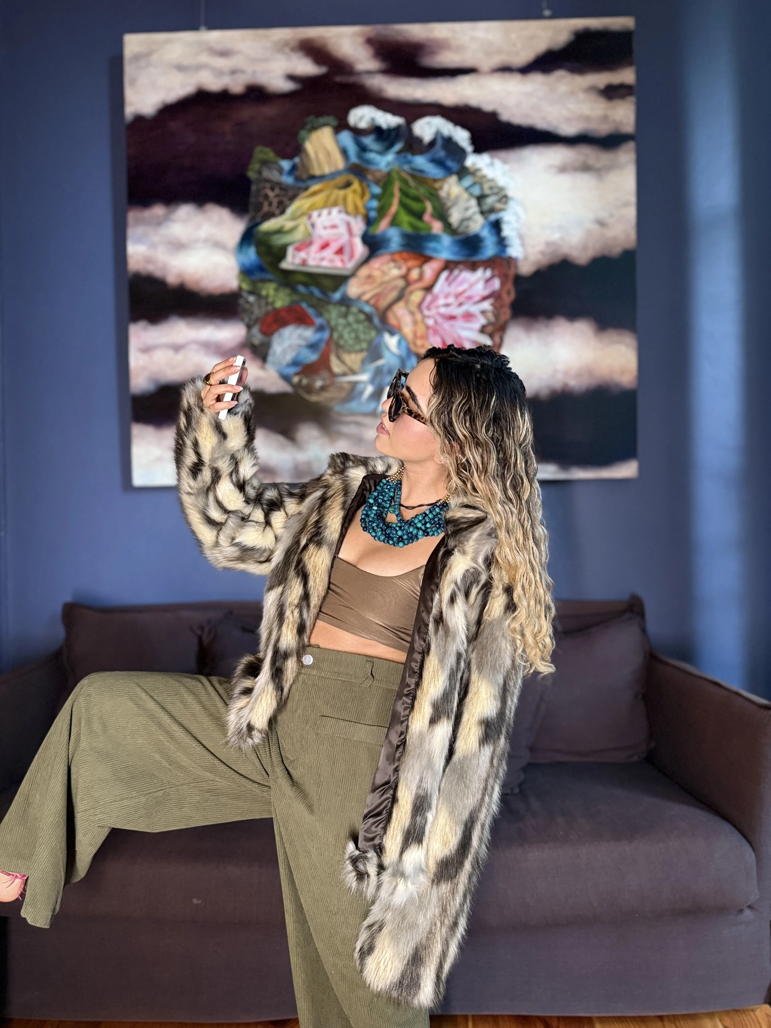 Una mujer con gafas, pelo rizado y maquillaje, usando un abrigo de piel y joyas, toma una selfie frente a una pintura colorida en una galería de arte.