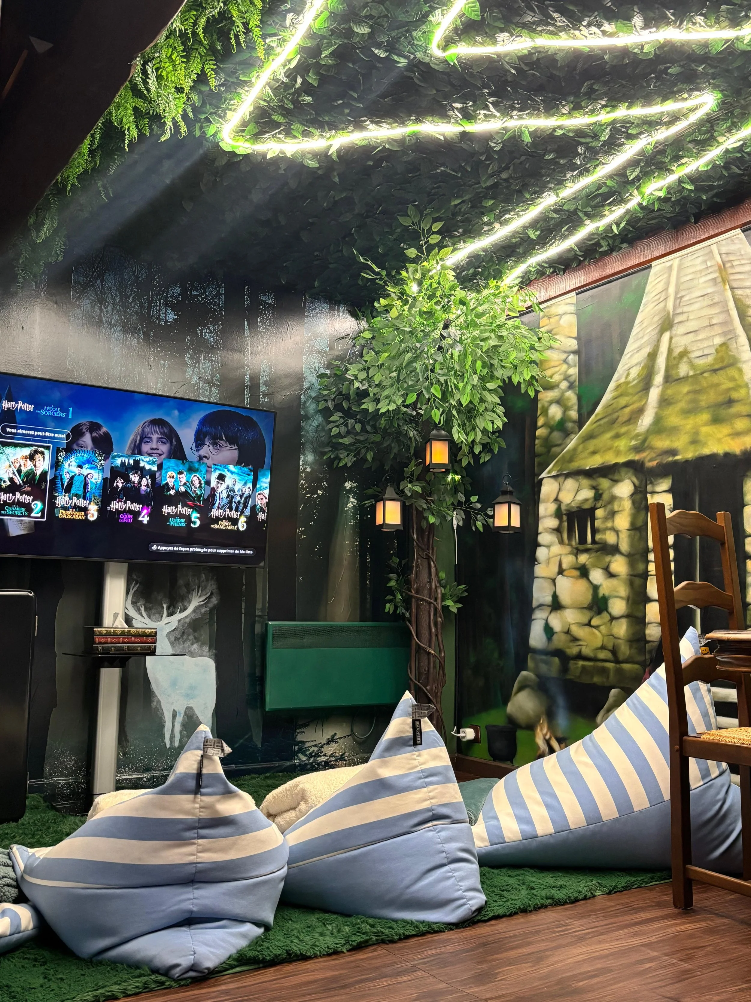 Salle avec décoration de forêt, plafond vert éclairé, poufs rayés, table en bois, mur peint avec une tour de pierre
