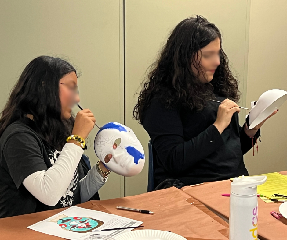 Two women painting white masks at a table during an arts and crafts activity.