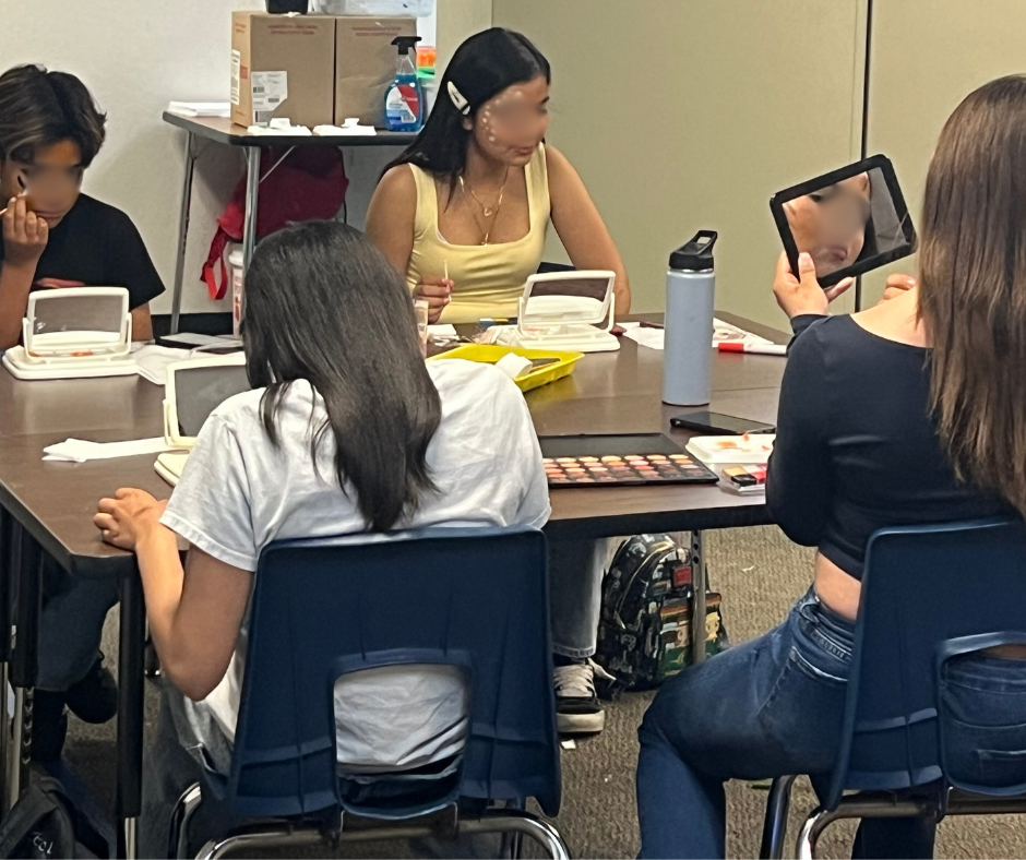 Group of women sitting around a table, applying makeup and using mirrors and a tablet