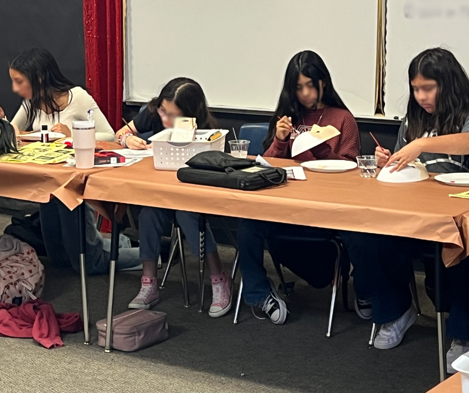 Four young girls sitting at a long table participating in a craft activity, with supplies like paper, scissors, and glue visible on the table.