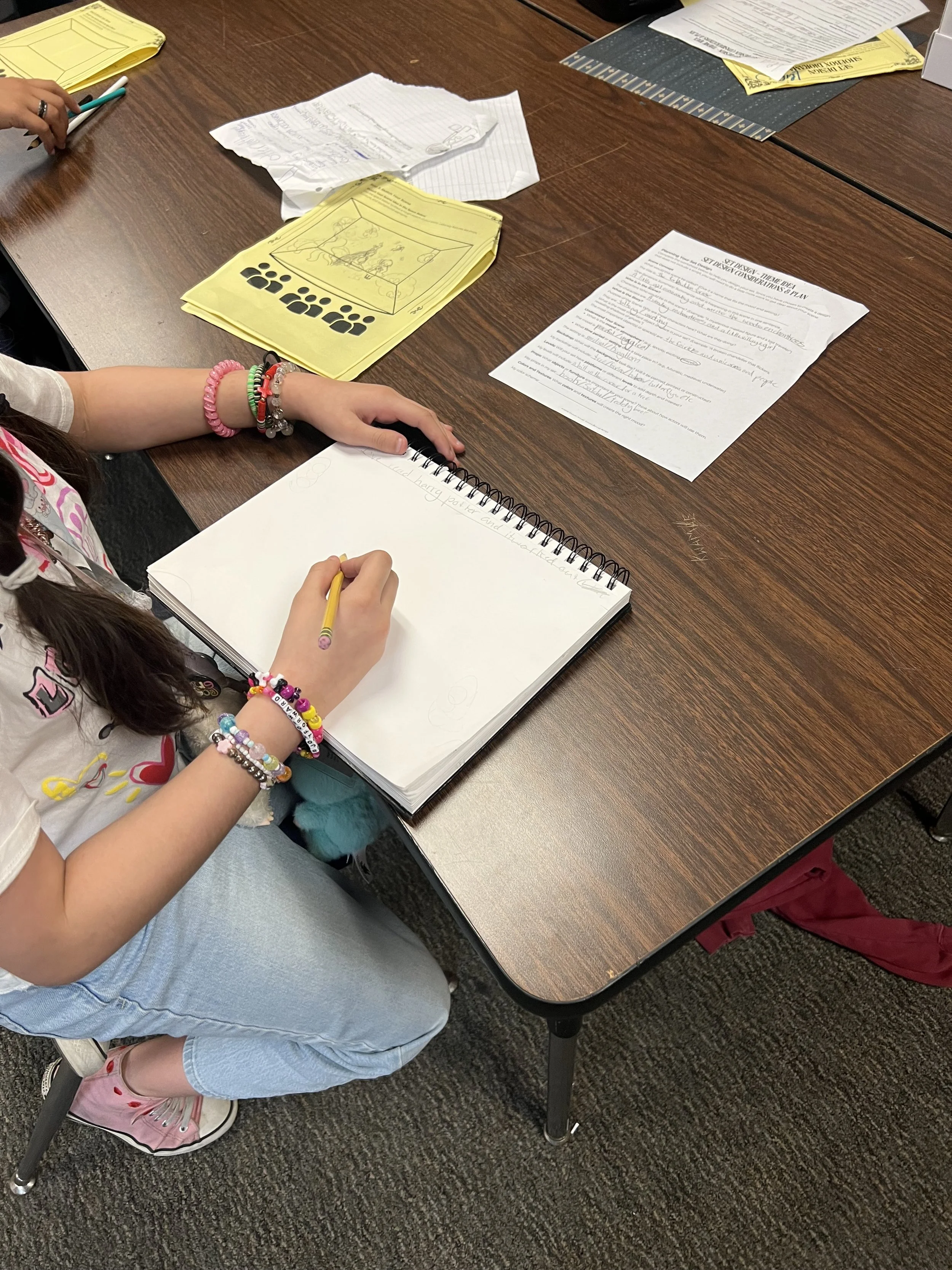 A young girl sitting at a wooden table, writing in a notebook with various papers and yellow colored papers scattered on the table.