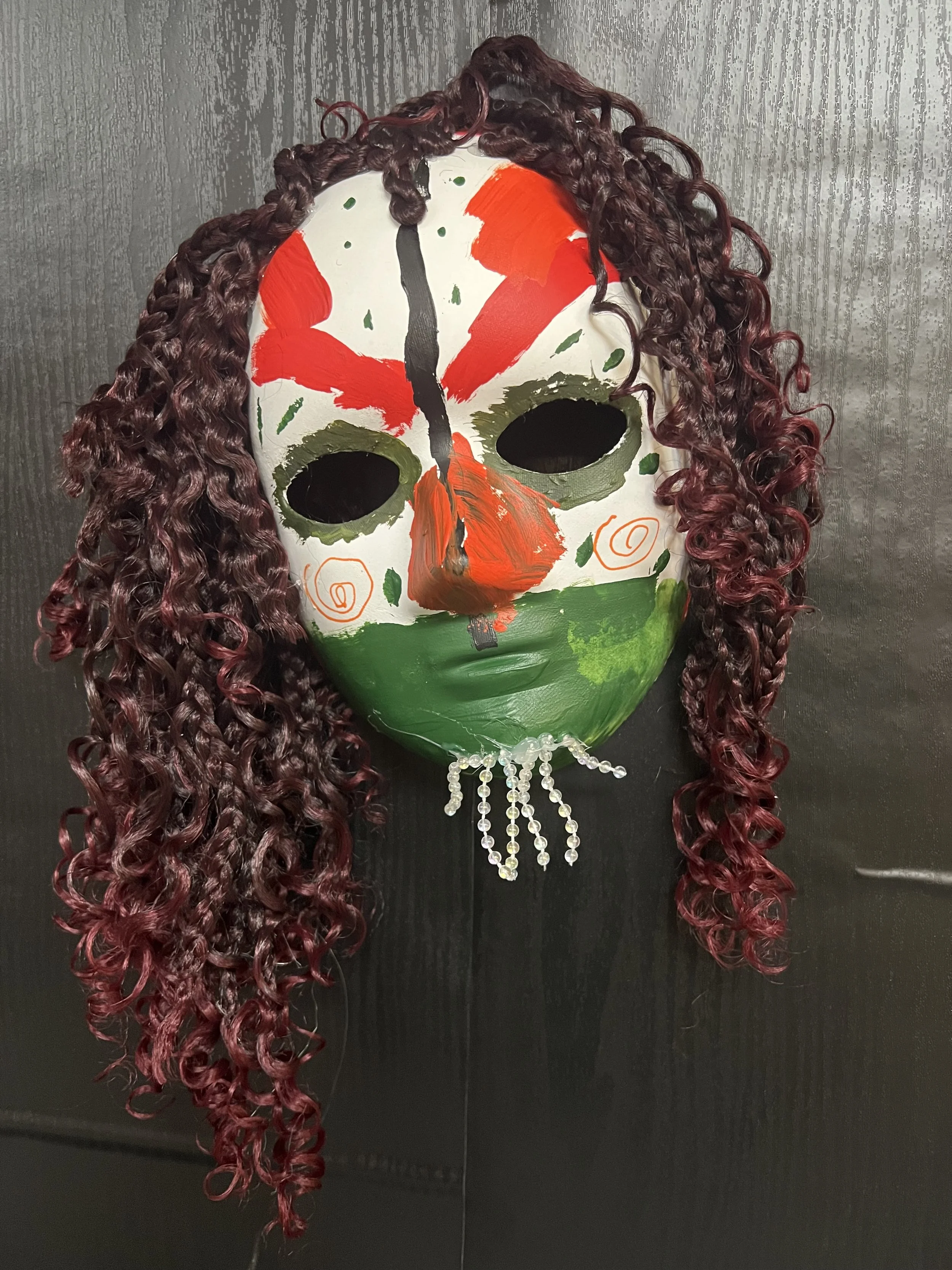 Decorative mask with painted face, curly burgundy hair, and rhinestone necklace, mounted on a black background.