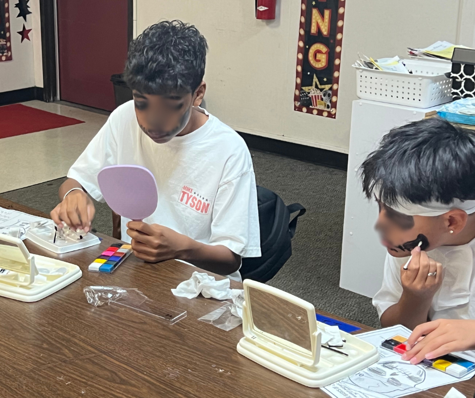 Two children sitting at a table, playing with makeup kits and small mirrors, one applying face paint and the other holding a small mirror, in a classroom setting.