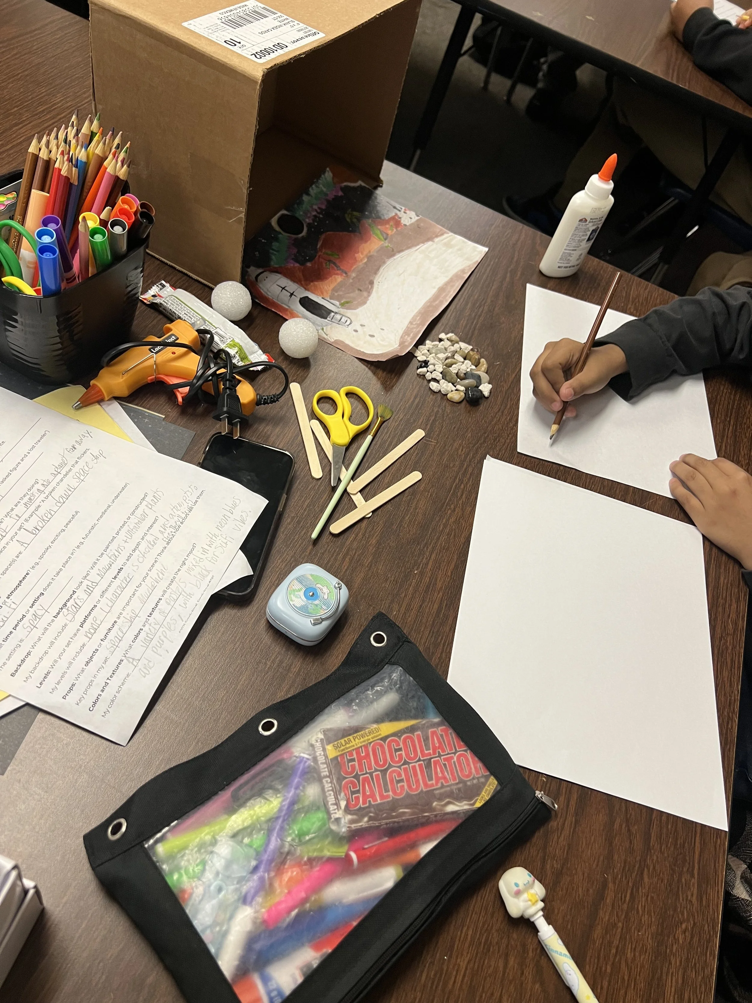 A cluttered craft table with colored pens, scissors, popsicle sticks, a glue bottle, rocks, a chocolate calculator, papers, and writing tools. Someone is drawing on a white sheet of paper.
