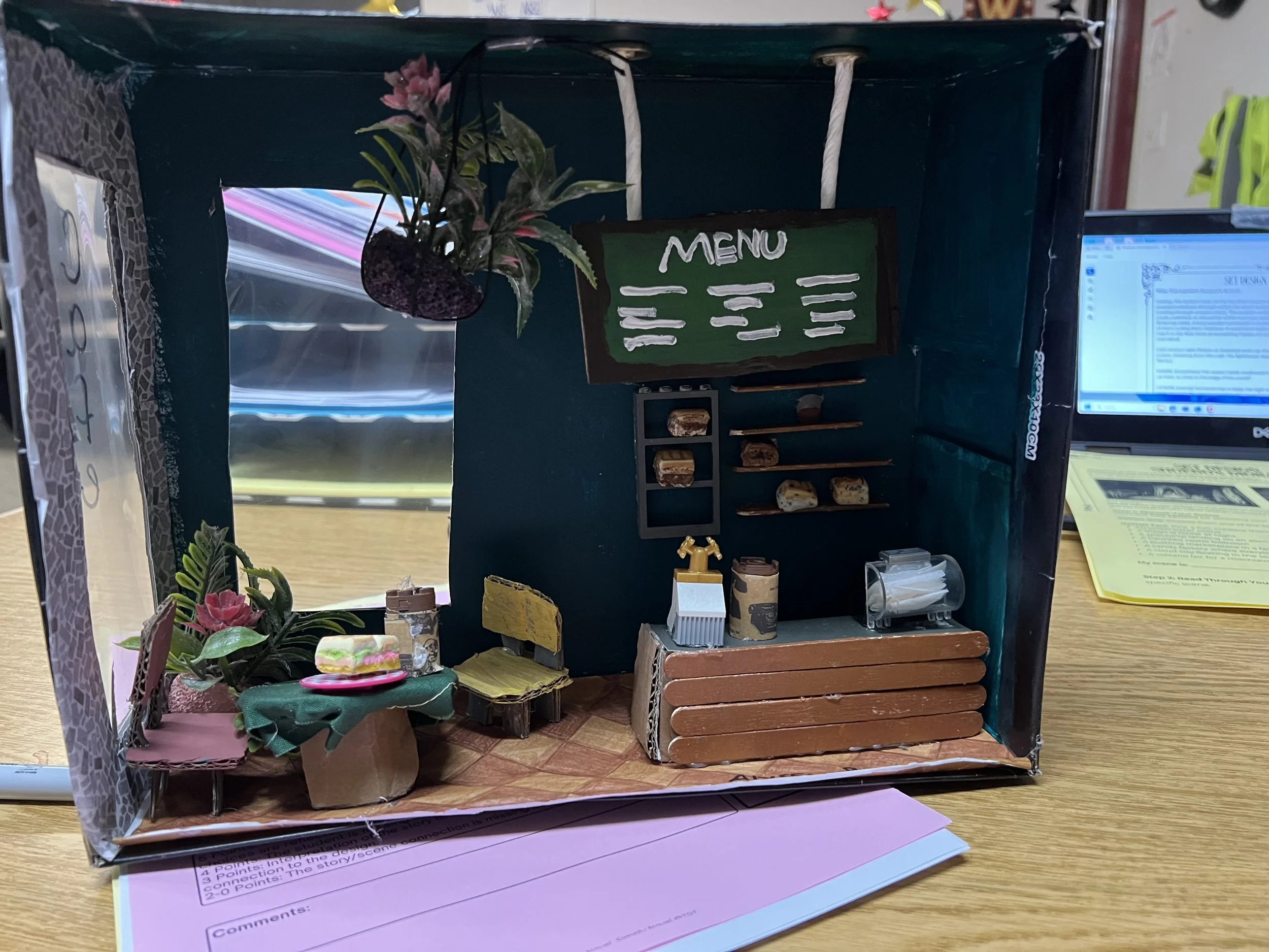 A miniature model of a cafe or restaurant with a green menu board, small furniture, and decorative plants, inside a cardboard box