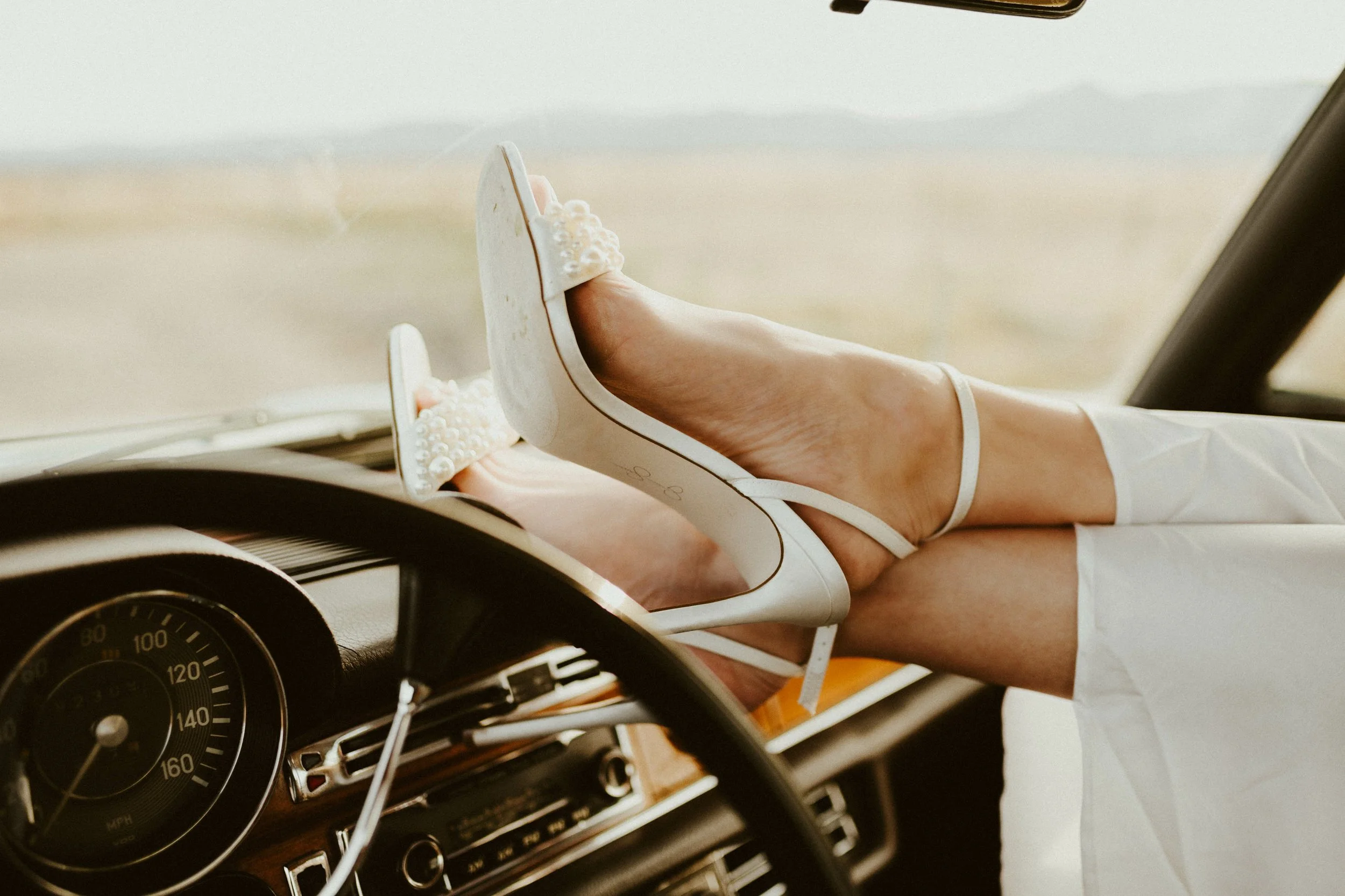 Close-up of a person lying in a vintage car, wearing white high-heeled shoes embellished with pearls, and white clothing, with landscape scenery in the background.