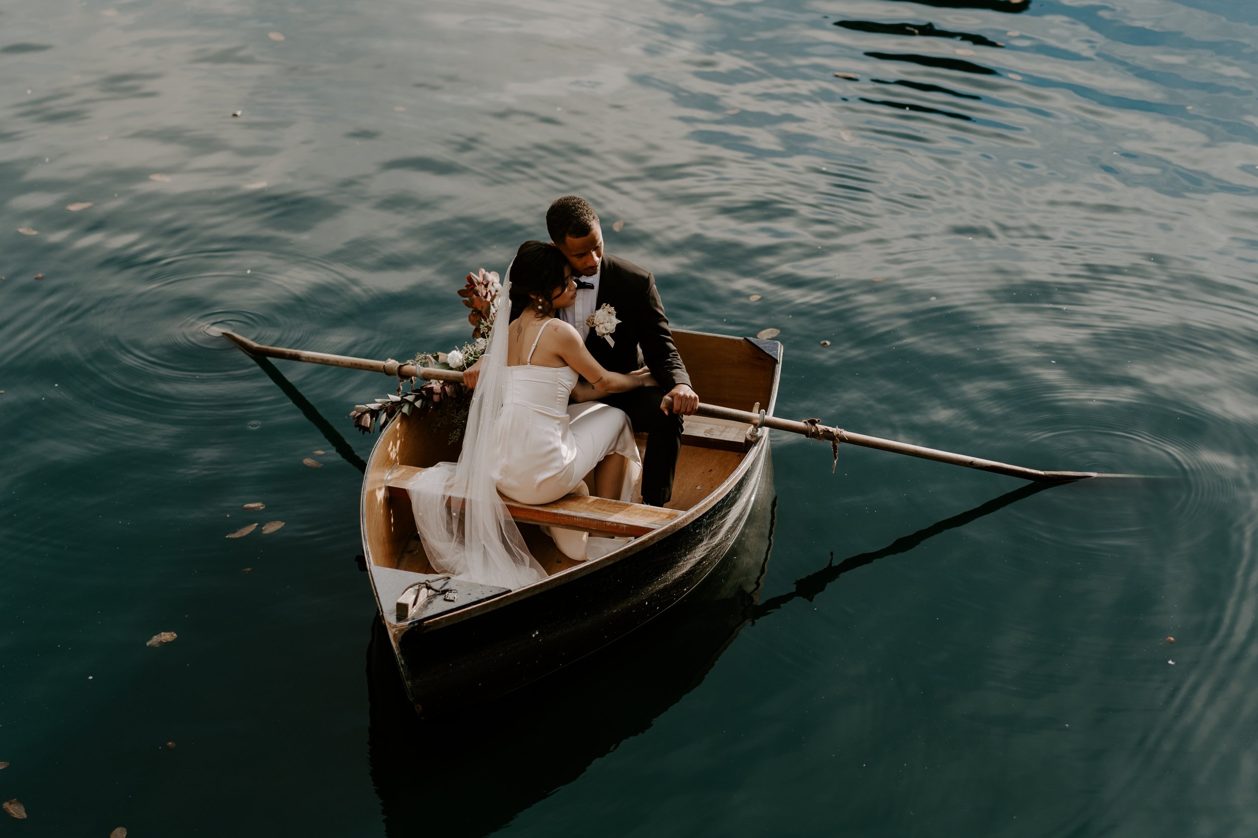 A bride and groom sitting together in a small boat on a calm body of water, embracing each other after their wedding, with the bride wearing a white dress and veil and the groom in a black suit.