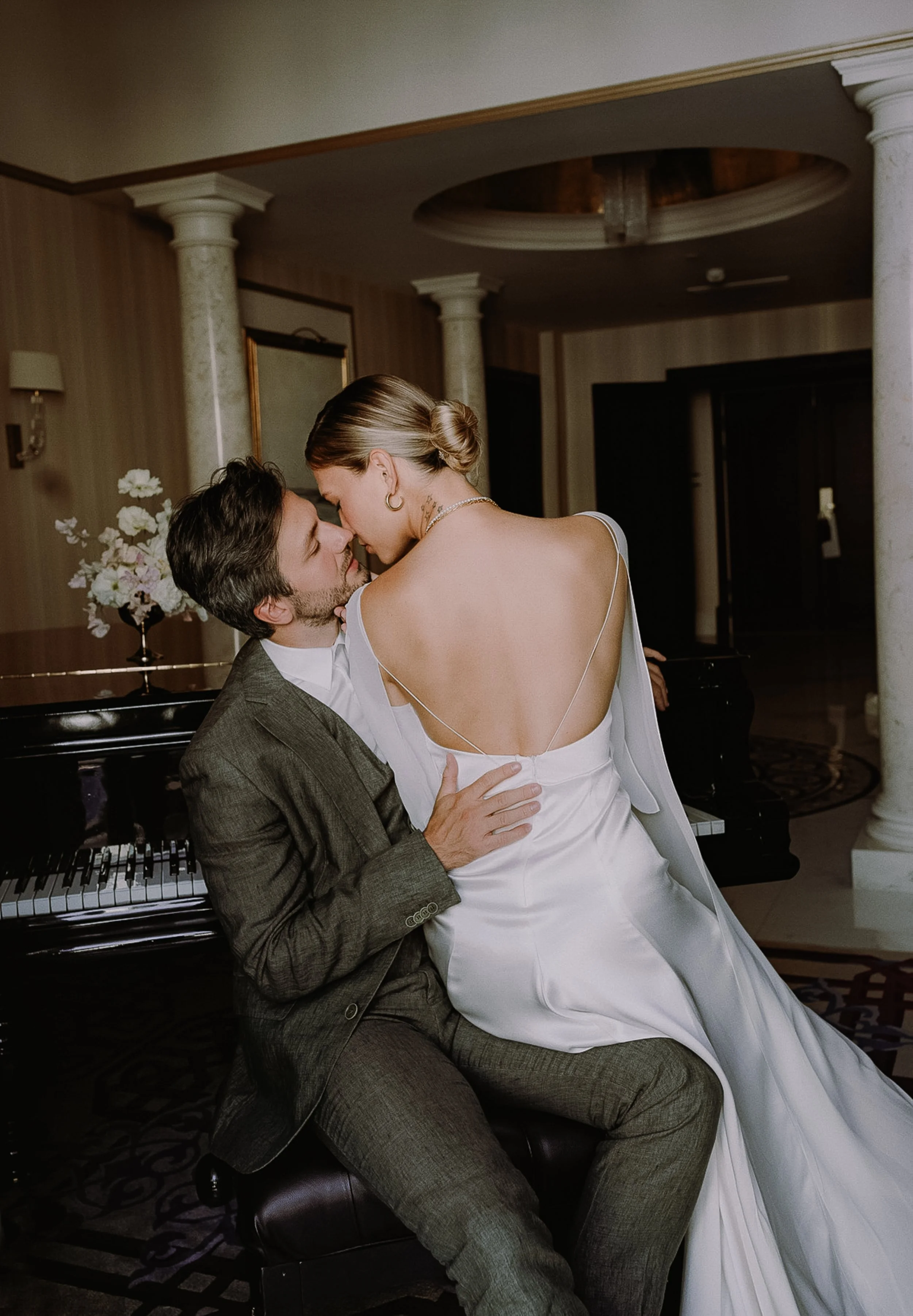 A couple sharing an intimate moment in an elegant room, with the woman dressed in a white gown and the man in a gray suit, near a black piano and floral arrangement.