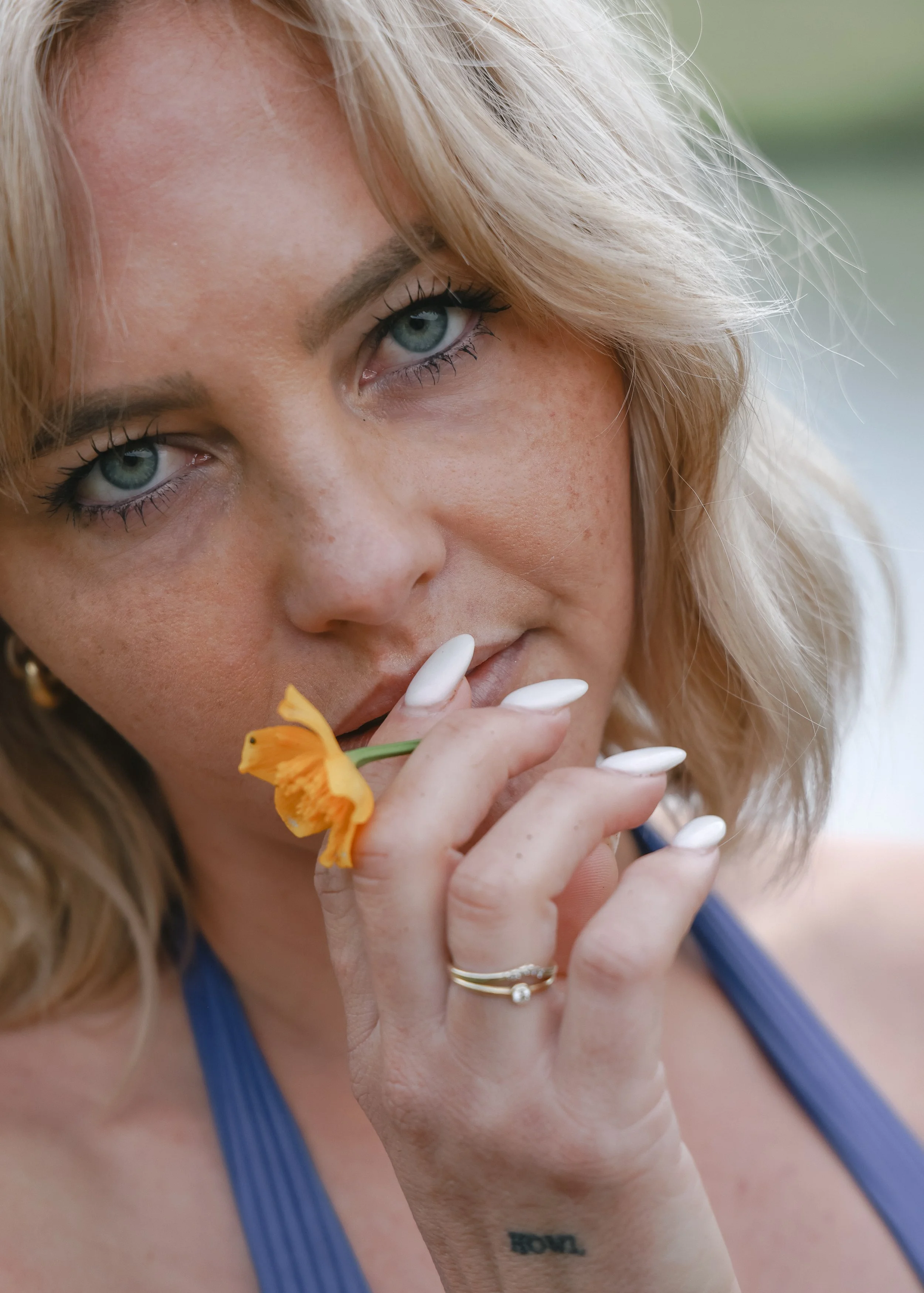 A woman with blue eyes and blonde hair holds a small orange flower close to her lips. She has white nail polish and wears a gold ring on her finger. She is wearing a blue sleeveless top.