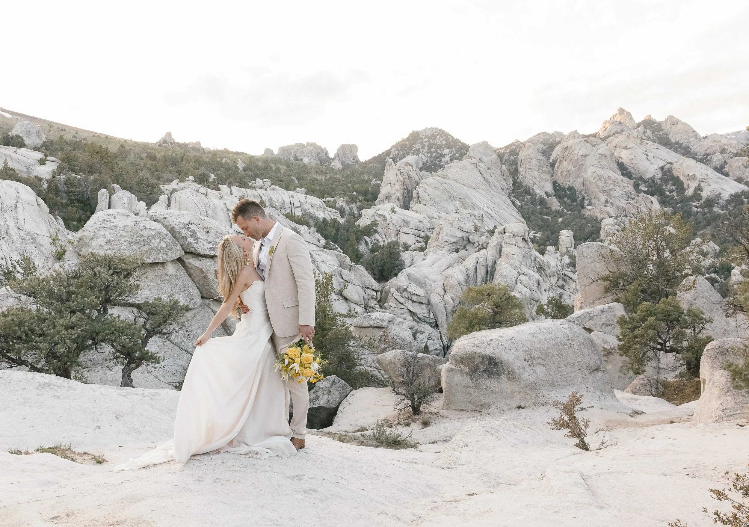 A bride and groom sharing a kiss in a rocky desert landscape with large boulders and mountains in the background during a wedding photoshoot.