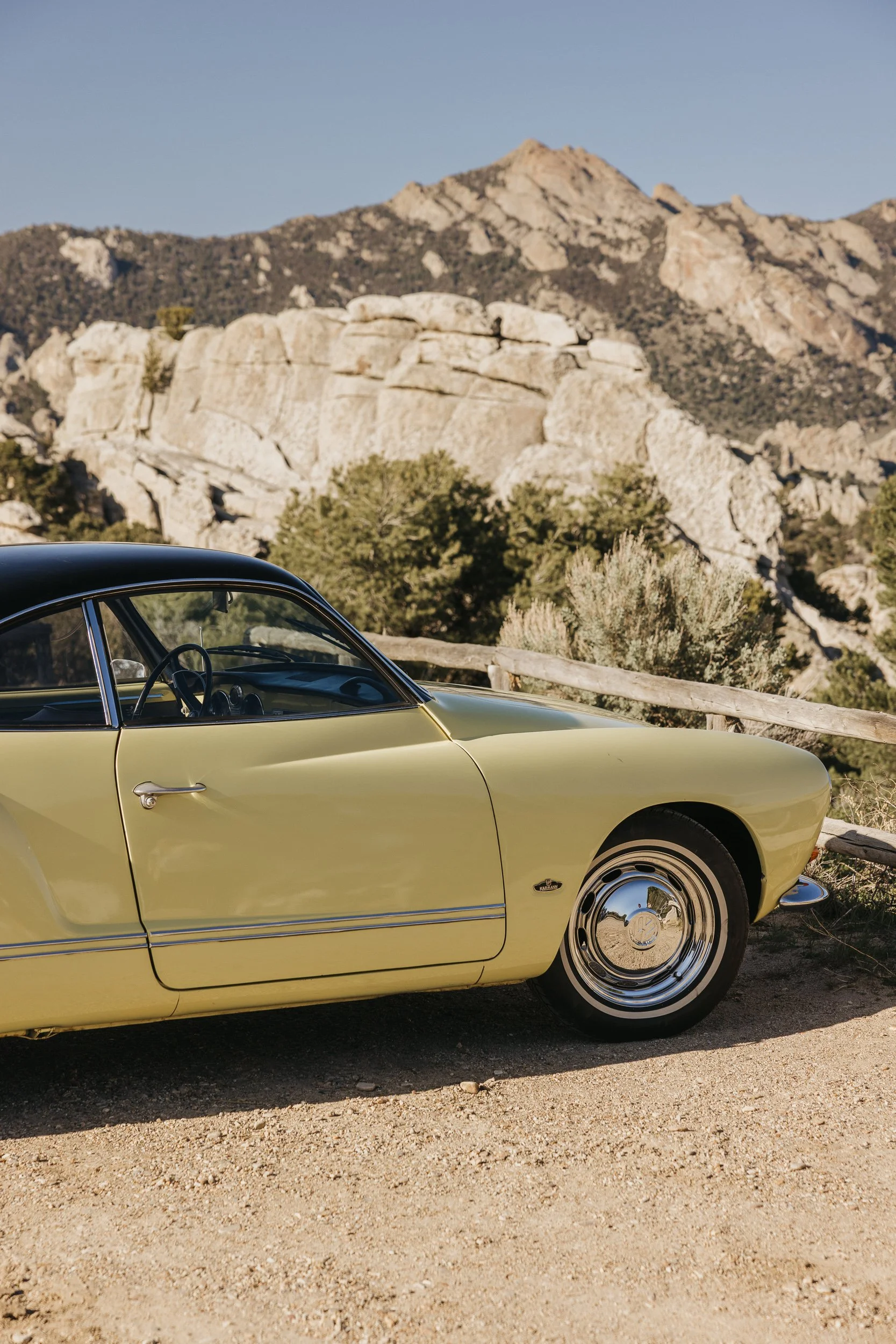 A vintage yellow car parked on a dirt road with rocky mountains and desert vegetation in the background.