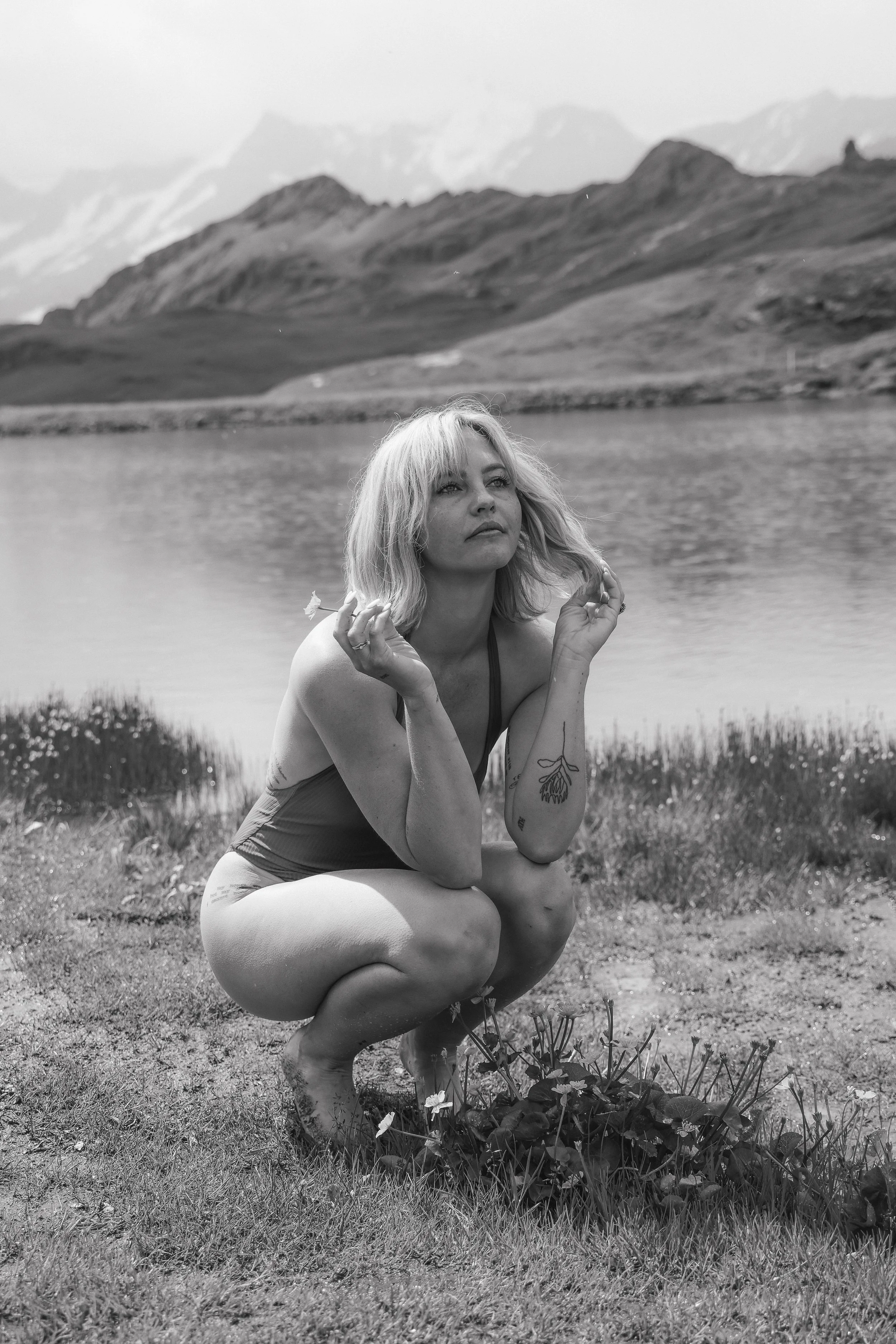 A woman crouching by a lake with mountains in the background, in a black-and-white photograph.