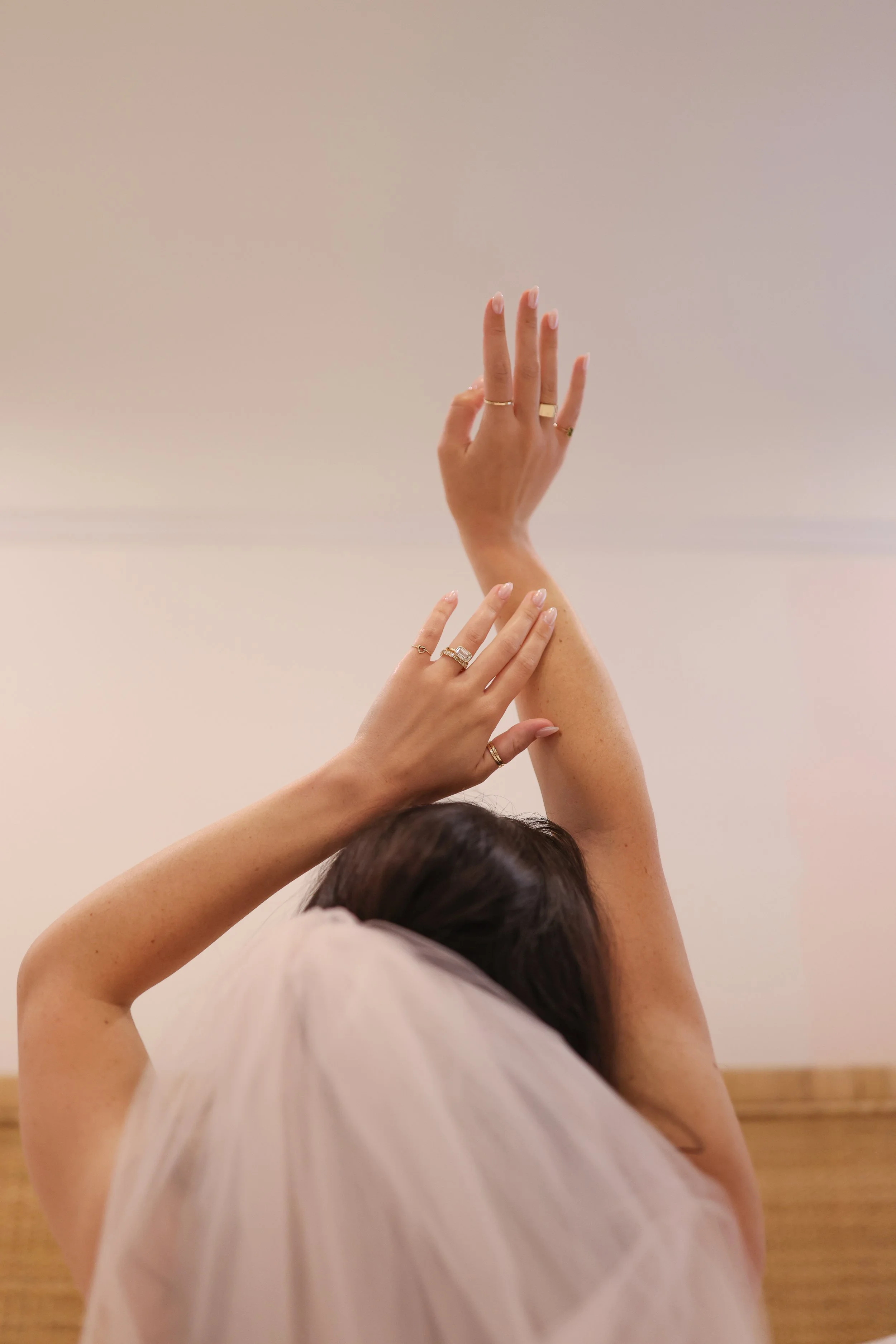 Woman wearing wedding rings with arms raised, one hand touching her head and the other extended upwards, against a plain background.